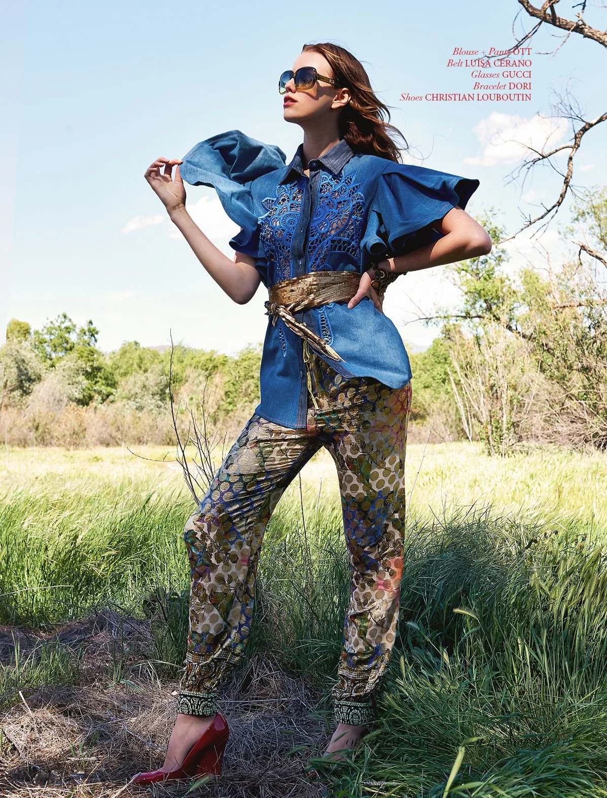 Full-length fashion shot by fashion photographer Daniella Hehmann in a rustic Los Angeles landscape featuring a model in a blue denim ruffle top and floral patterned trousers with an intricate gold waist belt.