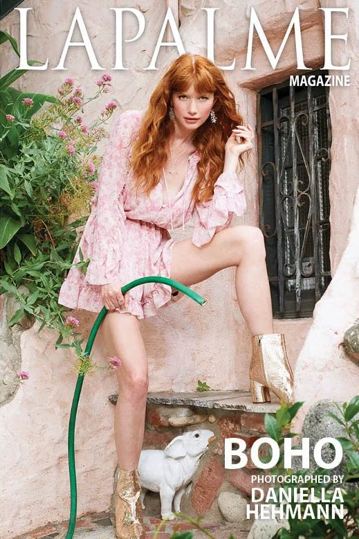 The cover of LaPalme magazine featuring a woman with long red hair in a pink dress, crouching on a ledge next to a white goat sculpture, holding a green garden hose, with a rustic stone wall and a window with iron bars behind her
