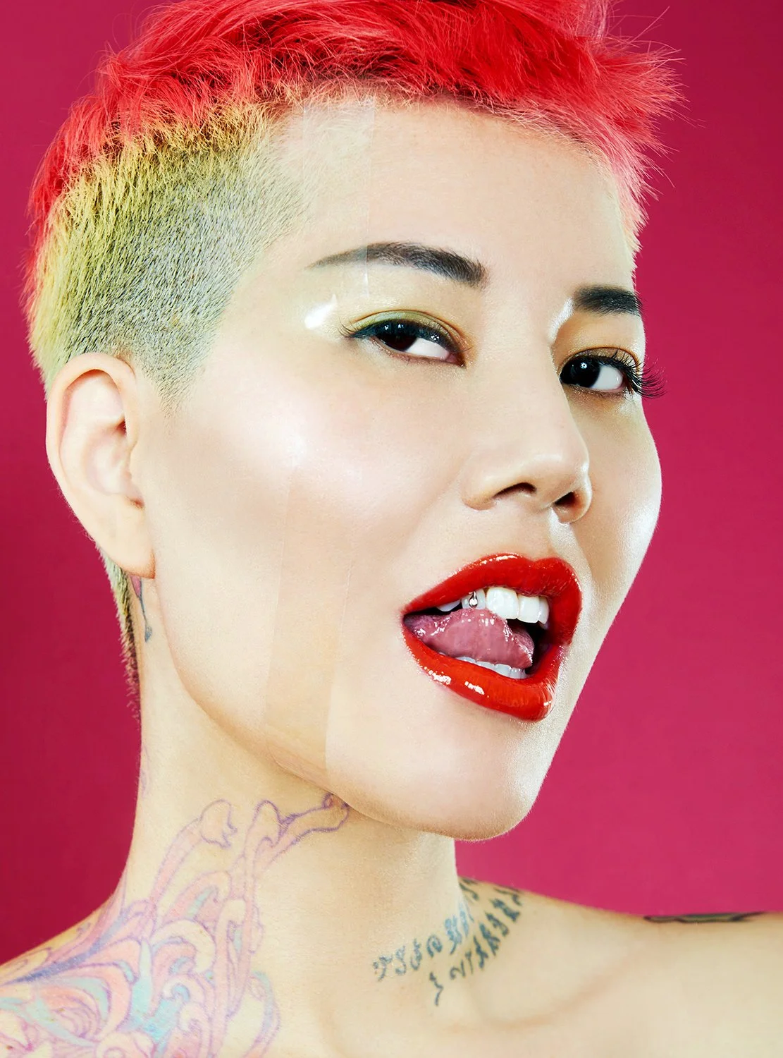 Close-up of a woman with short pink and blonde hair, red lipstick, and tattoos on her neck and shoulder, posing with her tongue out slightly and a playful expression against a pink background.