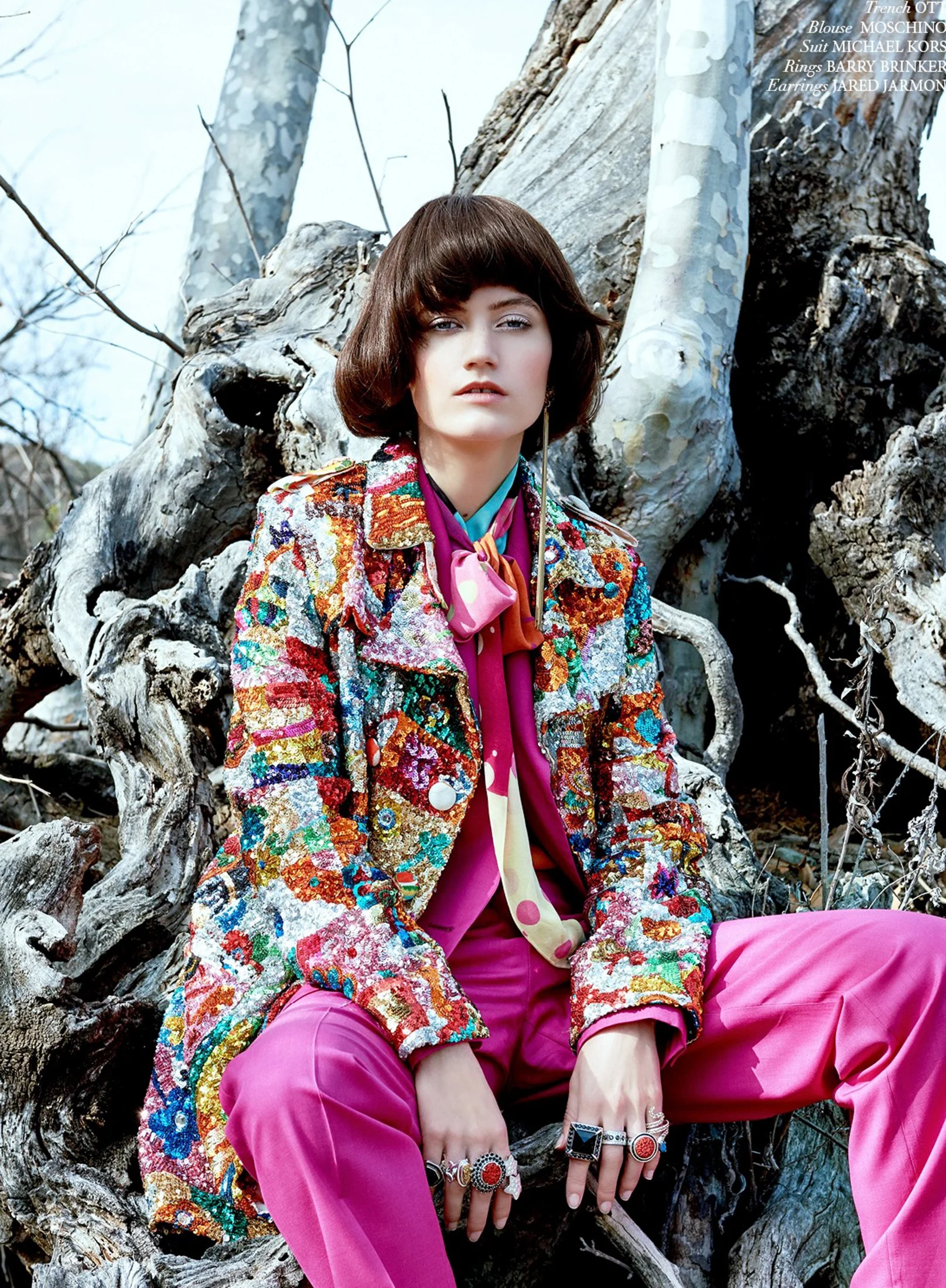 A vivid fashion portrait by Daniella Hehmann Photography shot in L.A. featuring a model in a heavily patterned, colorful floral coat and bright pink trousers, seated against a rugged, natural outdoor backdrop. She has multiple rings on her fingers an