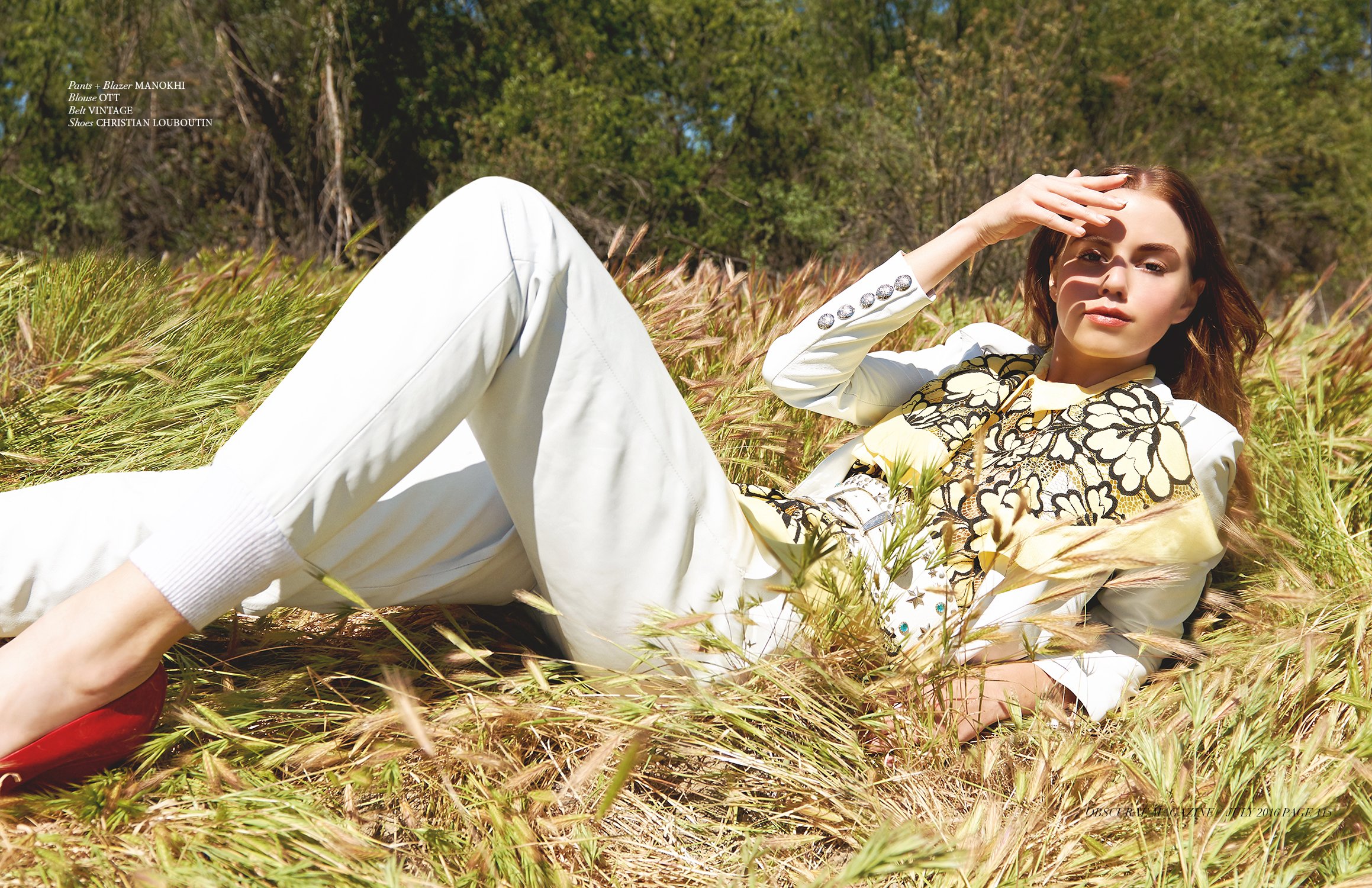 A woman lying in a field of tall grass with trees in the background, dressed in white pants and a patterned jacket. She is looking at the camera while shielding her eyes from the sun.