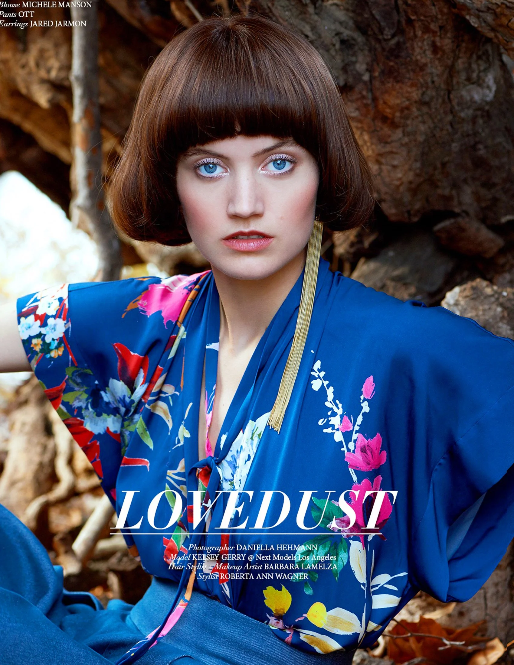 Cinematic editorial photography by photographer Daniella Hehmann in Los Angeles. A model with a bob haircut poses in a deep blue floral kimono-style top with 'LOVEDUST' text overlay, set against warm-toned rocks.