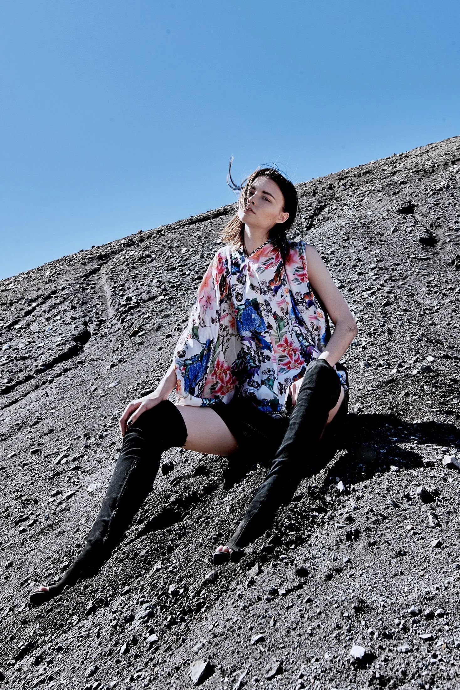 A woman sitting on a rocky, sloped surface under a clear blue sky, wearing a floral sleeveless top and black thigh-high boots.