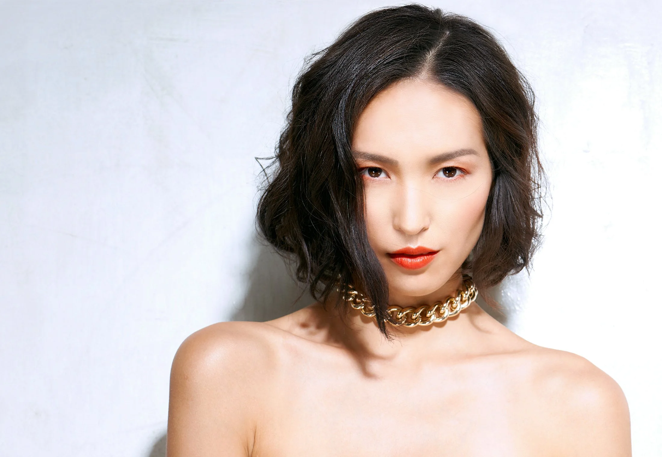 A woman with wavy short black hair wearing a gold chain necklace, red lipstick, and natural makeup, looking directly at the camera against a plain white background.