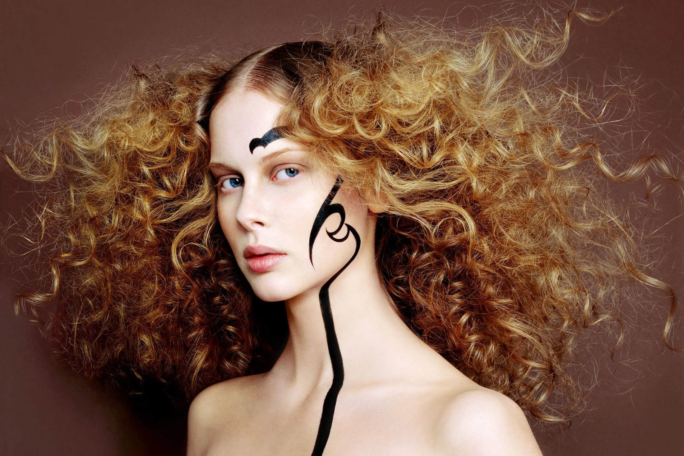 A woman with long, curly red hair and blue eyes has black lines and shapes drawn on her face and neck, against a brown background.