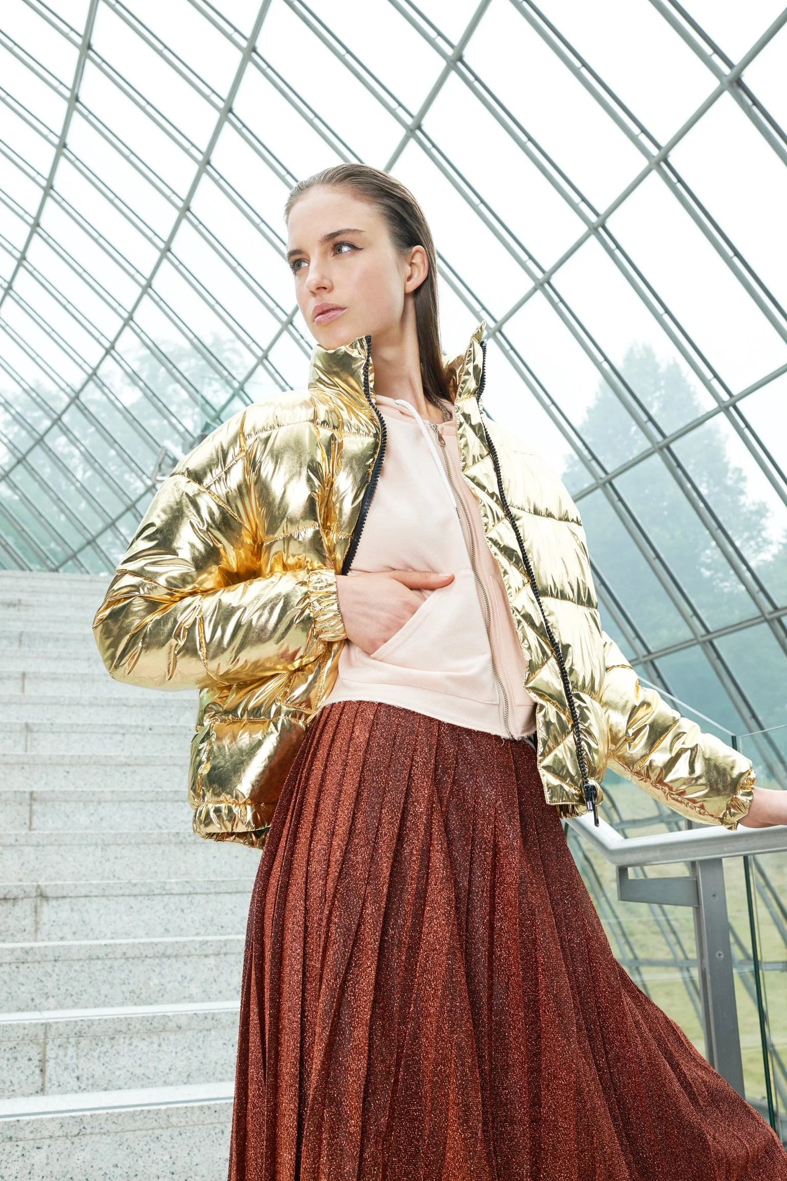 A woman wearing a shiny gold puffer jacket, a light-colored hoodie, and a long, pleated, reddish-brown skirt standing indoors near a large glass structure with stairs in the background.