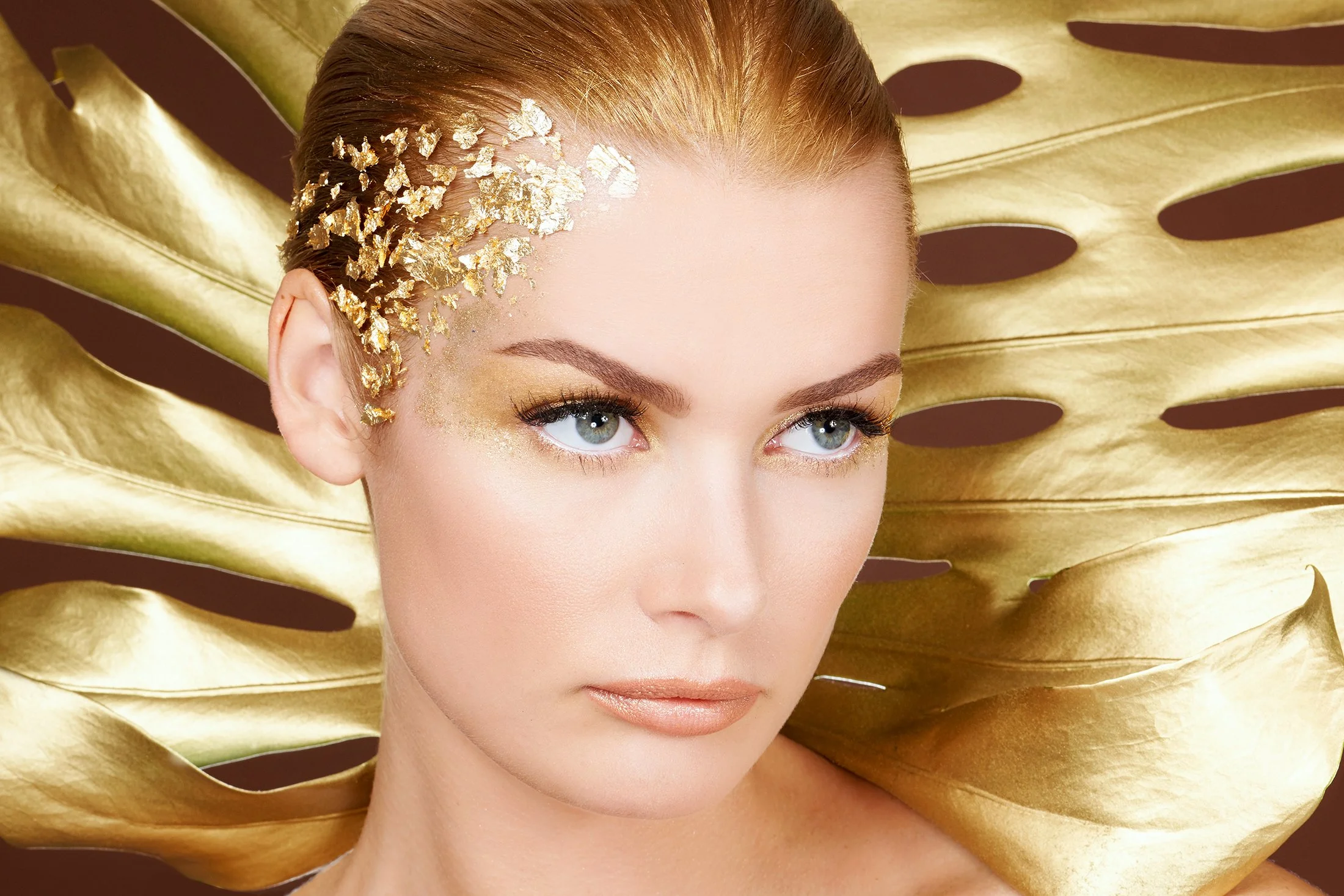 Professional beauty photography for Senna Cosmetics by Daniella Hehmann, showcasing makeup by Eugenia Weston. The editorial lighting is crisp, highlighting the intricate gold leaf hair accents and golden eye makeup. A large, perforated metallic gold 