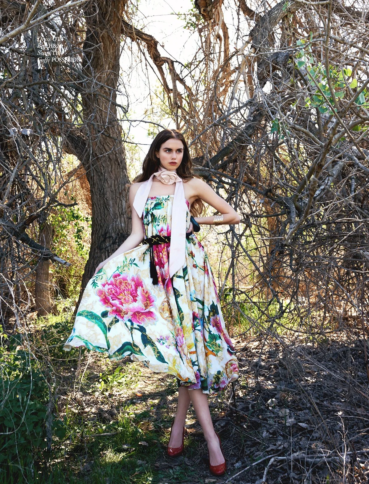A woman in a floral dress poses outdoors among trees and branches, wearing red high heels and a scarf around her neck.