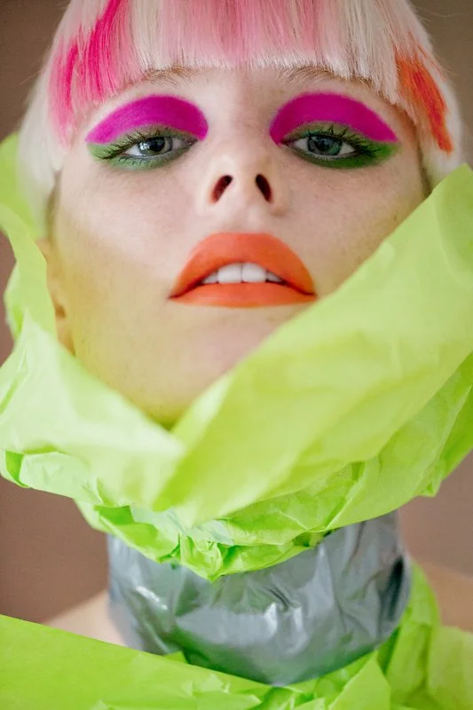 A female model with colorful makeup, pink hair, bright orange lipstick, and green eye shadow, wrapped in green and silver tissue paper.