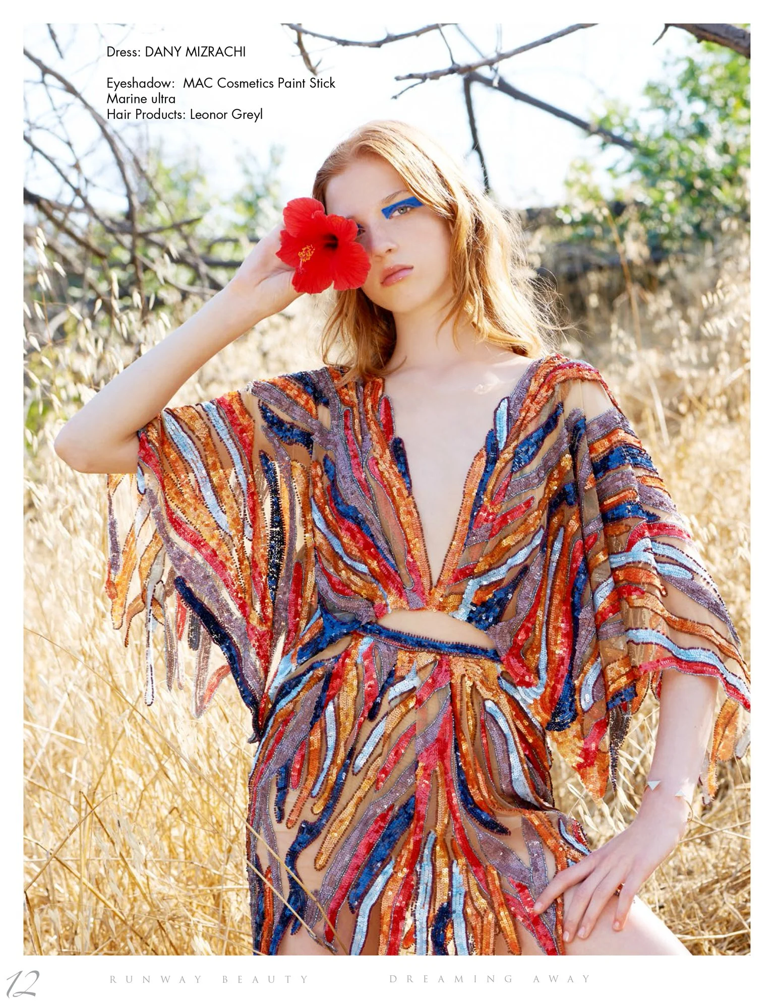 Model standing in a golden field wearing a colorful, sequin-embellished dress with a deep V neckline and cutouts, holding a red flower to her face with one hand, with natural background and trees