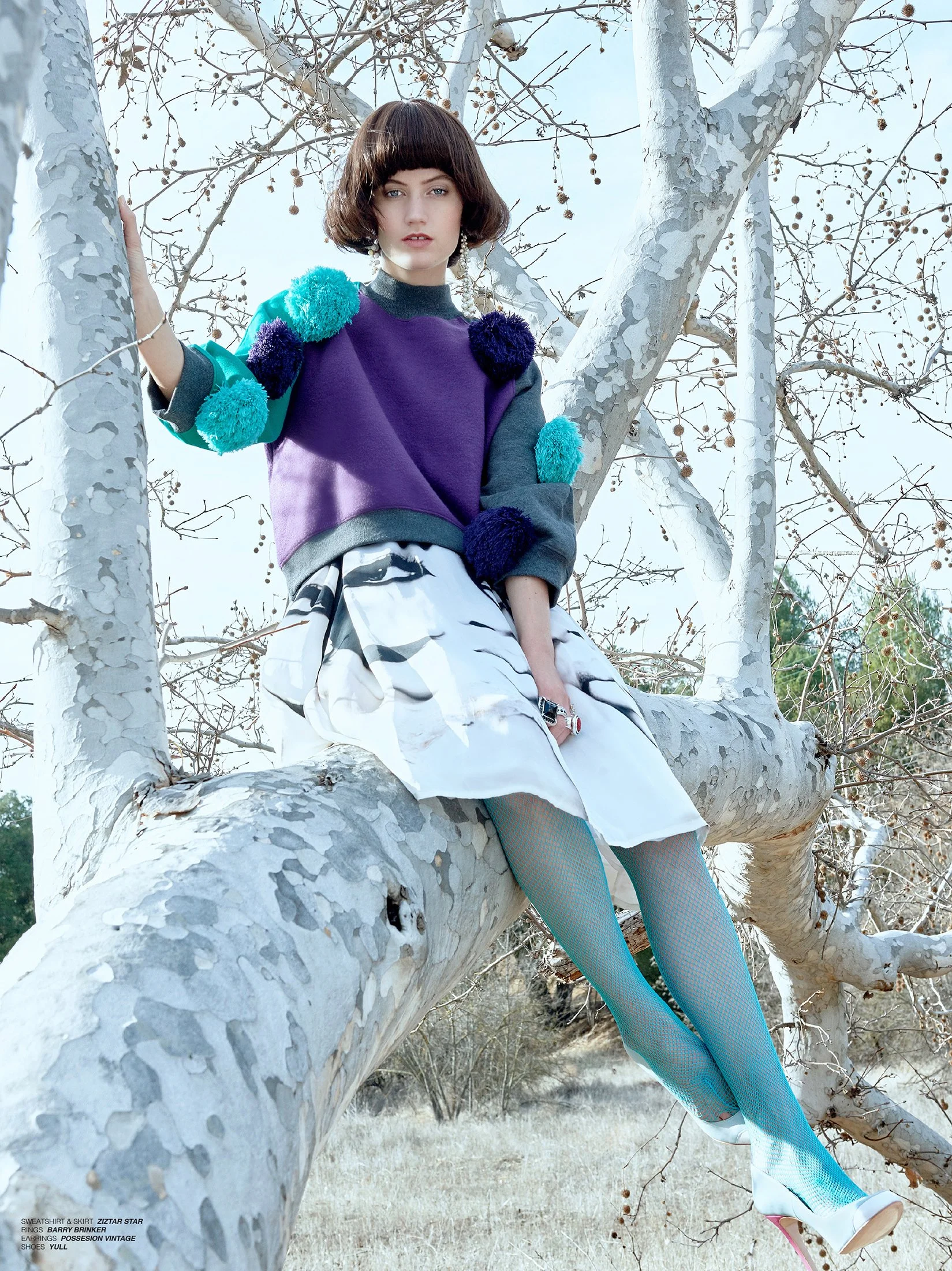 High-fashion outdoor shot by Daniella Hehmann Photography in L.A. featuring a model in a colorful sweater with pom-poms, a white patterned skirt, fishnet tights, and white shoes with pink soles. The background shows a clear sky and leafless trees.