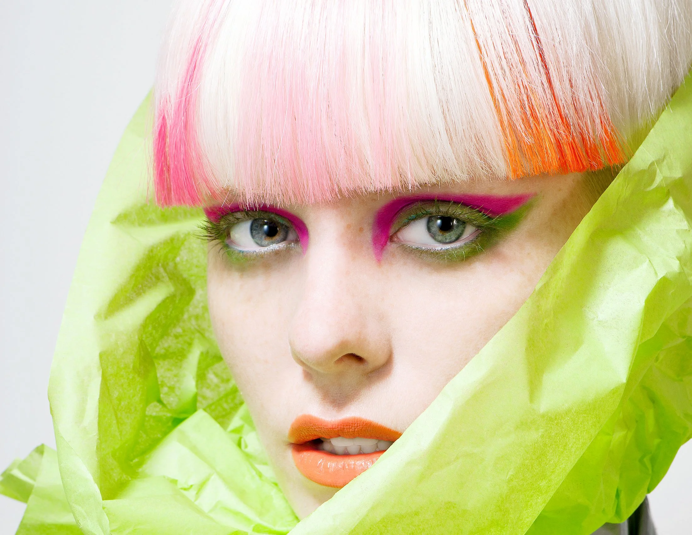Fotografie von Daniella Hehmann: A model with pink and blonde hair styled in a short bob with straight bangs, surrounded by green tissue paper, wearing vibrant pink and green eye makeup, peach lipstick, and gazing directly at the camera. 