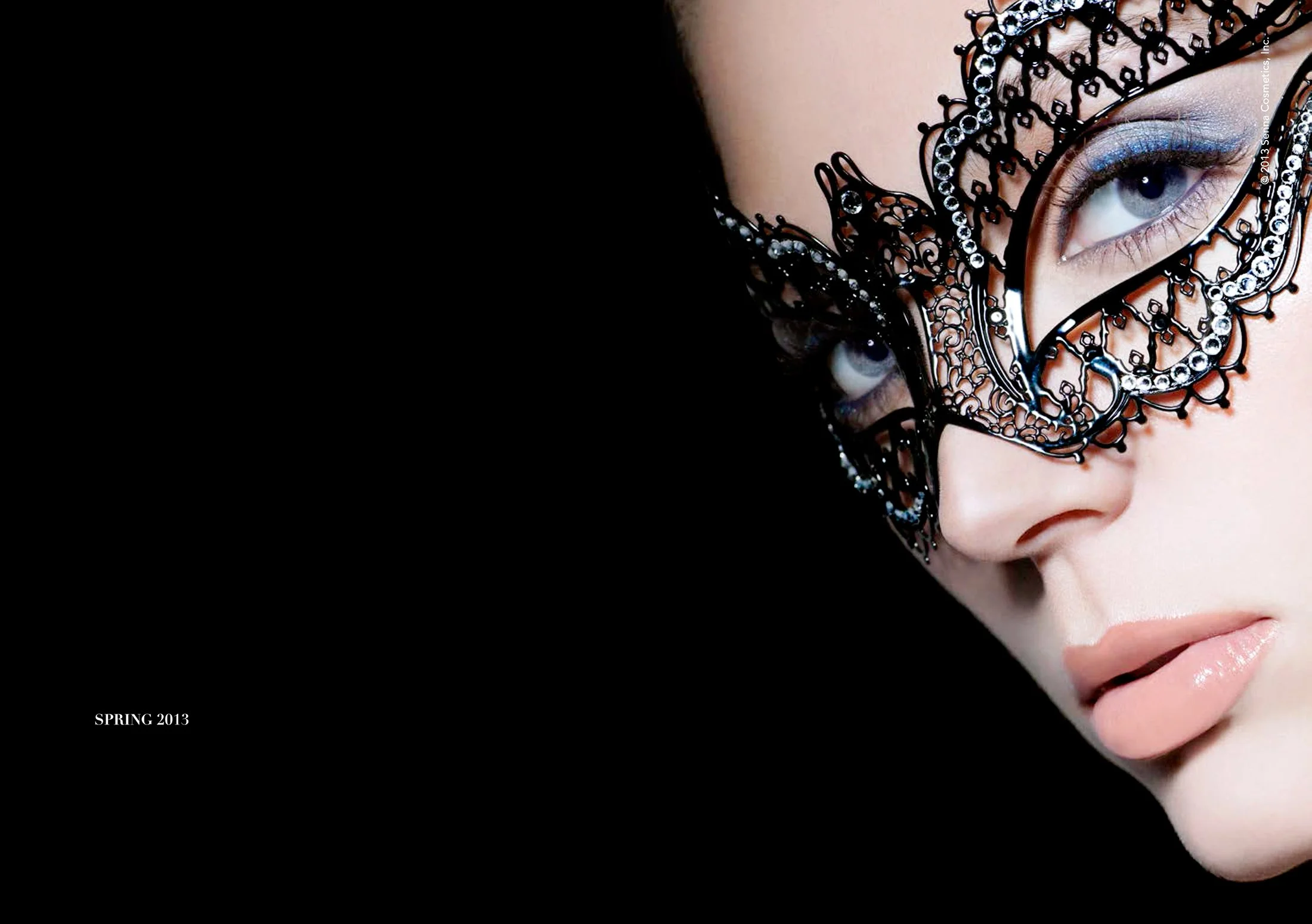 Close-up image of a woman wearing an ornate black mask with rhinestones, highlighting her blue eye and pink lips, with a black background. Text reads 'SPRING 2013' in the bottom left corner.