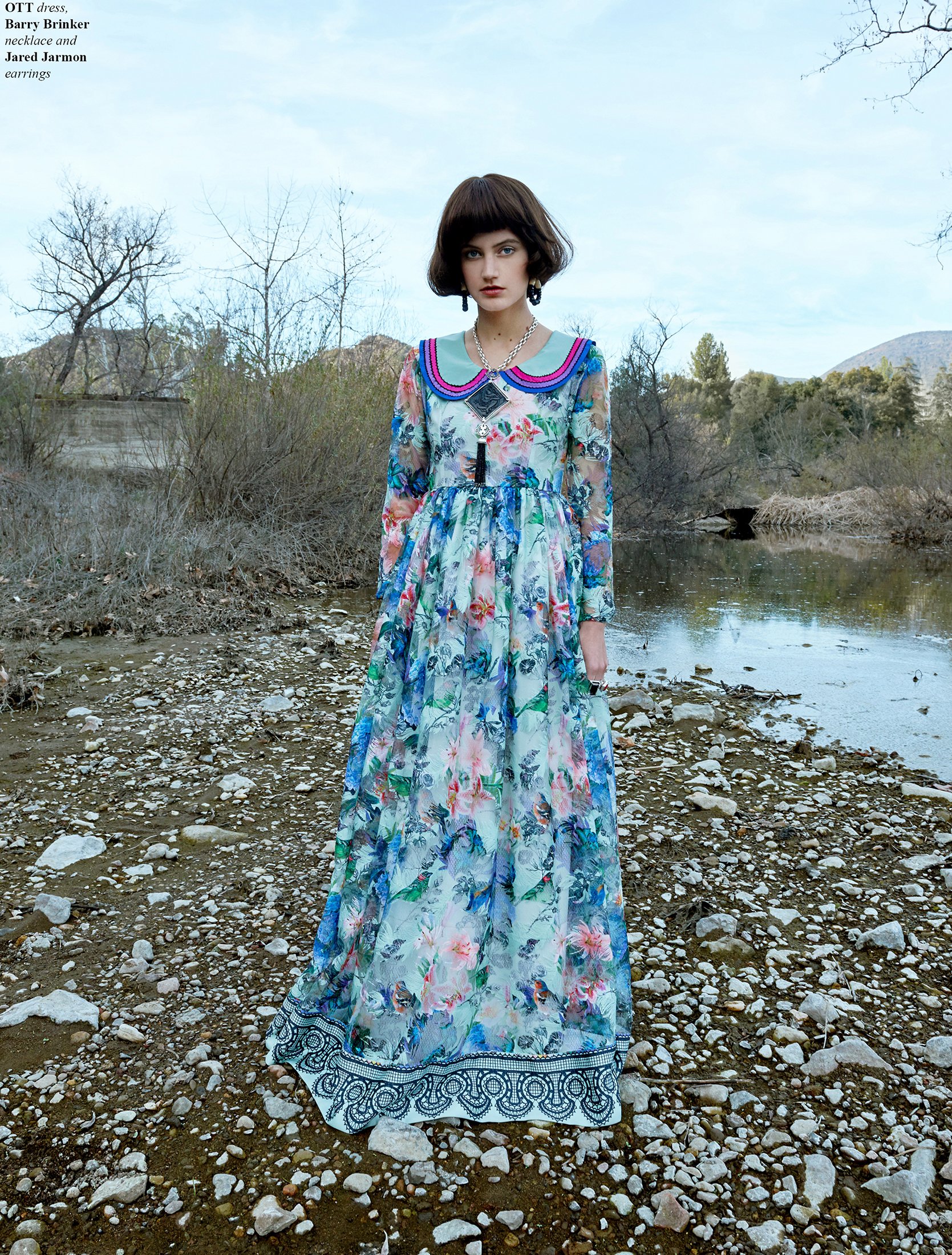A full-length fashion shot by Daniella Hehmann Photography in L.A., featuring a model in a floor-length floral gown standing before a serene lake with dry winter foliage in the background.