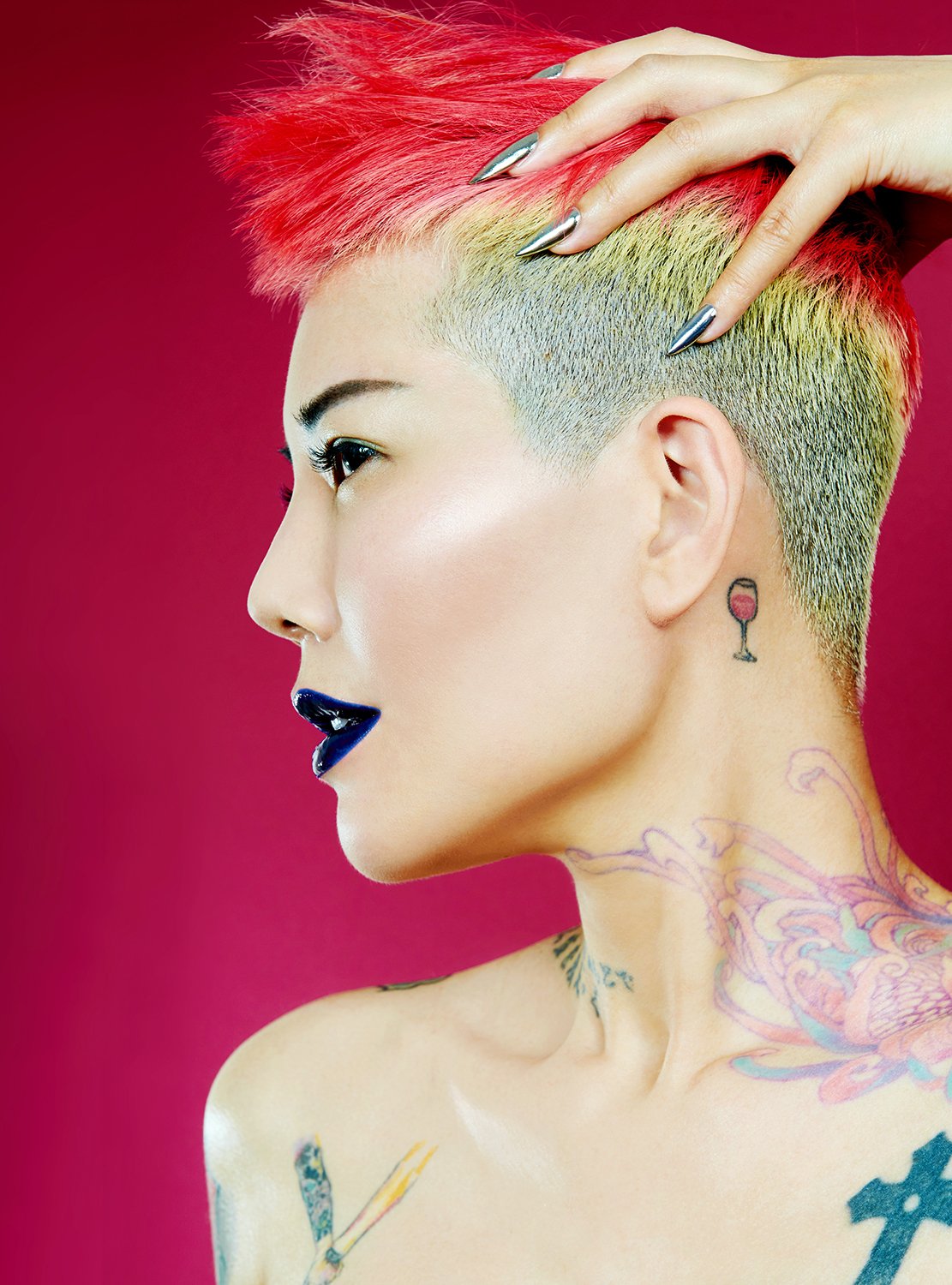 A woman with vibrant pink and yellow short hair, tattoos on her neck and shoulder, and bold makeup including dark lipstick, posing against a pink background.