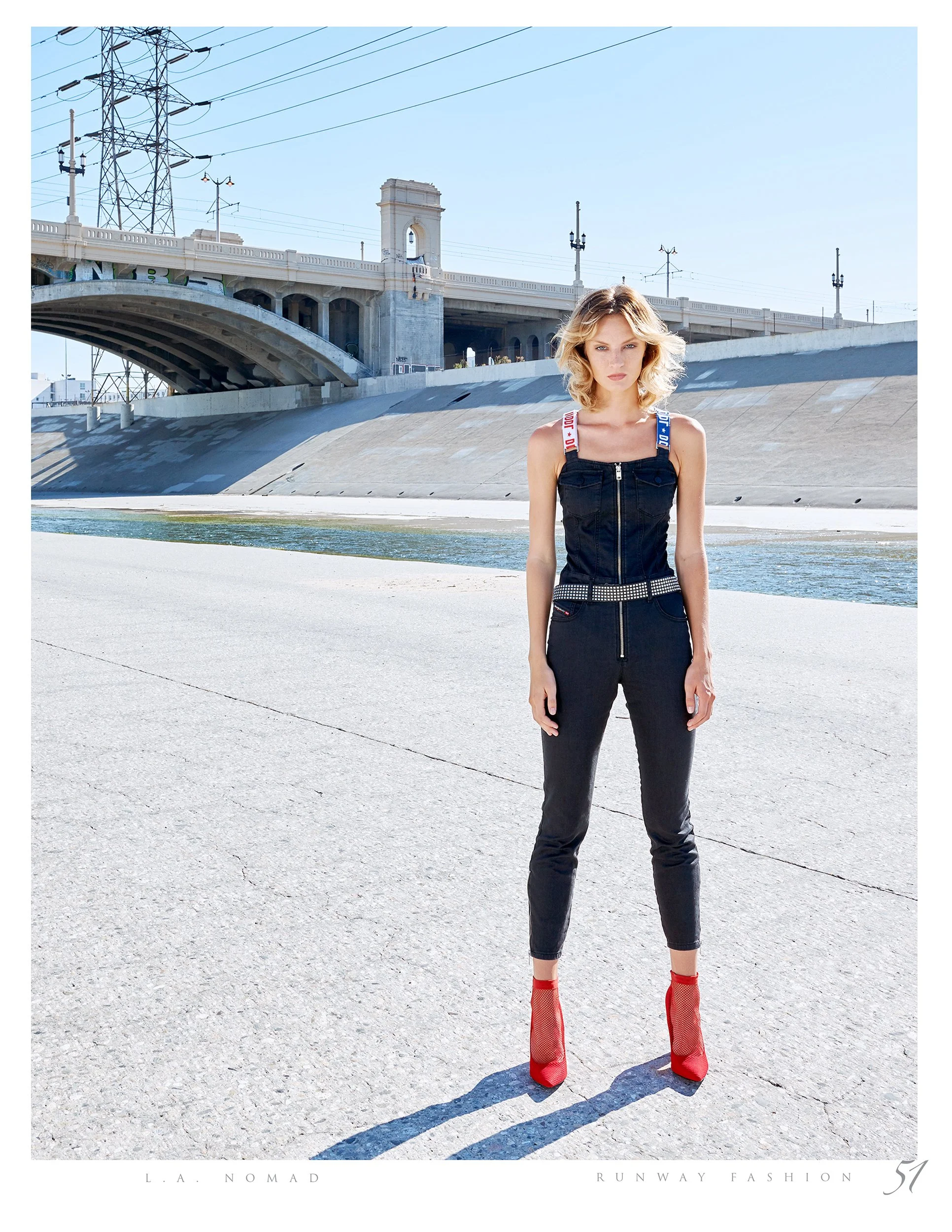 A woman standing on a concrete surface near a waterway, wearing a black denim jumpsuit, red high heel shoes, and an embellished belt, with a bridge and power lines in the background.