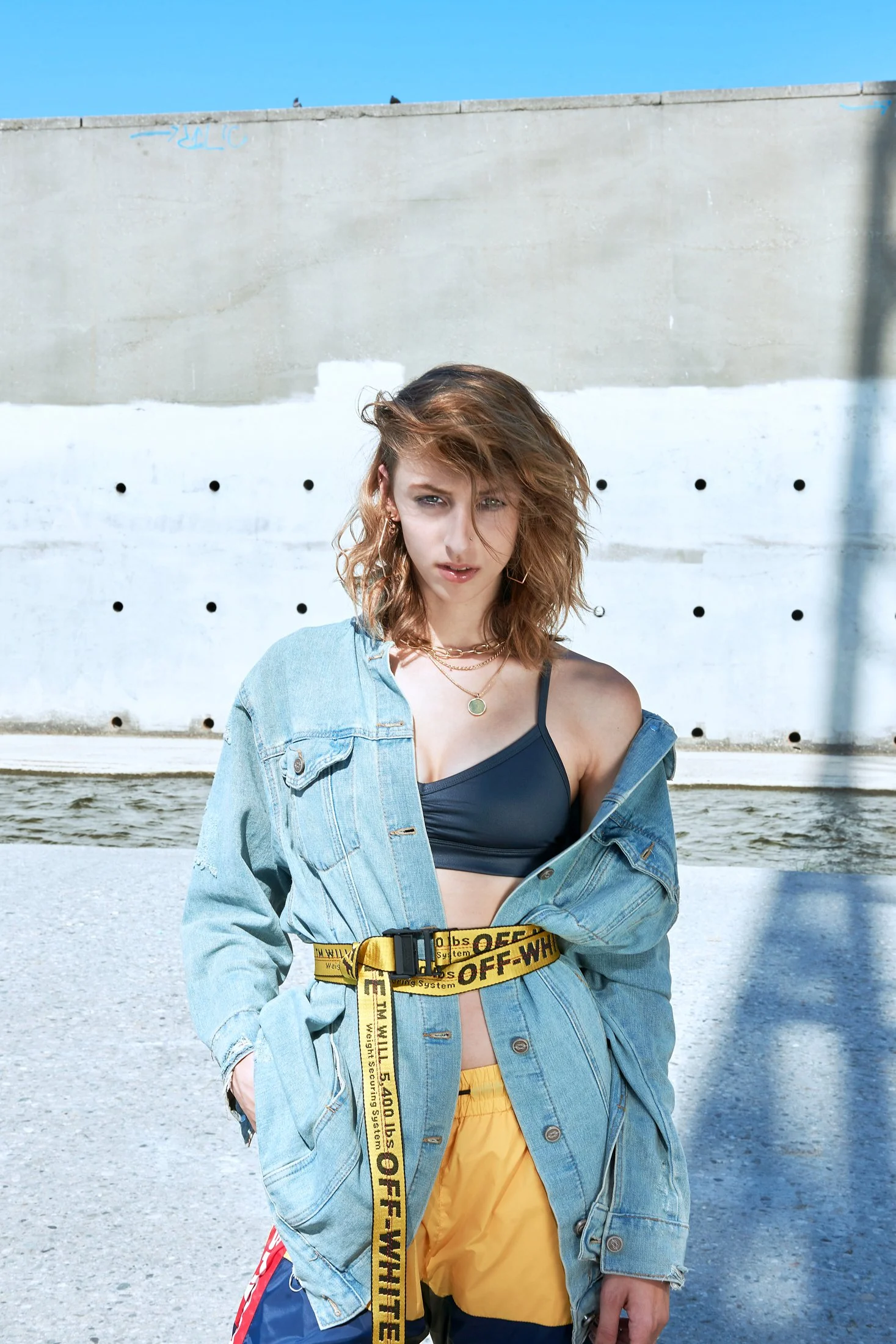 A high-contrast fashion shot by photographer Daniella Hehmann in L.A. featuring a model in a denim jacket with a bold yellow 'Off-White' style belt, captured against the industrial backdrop of the Los Angeles River.