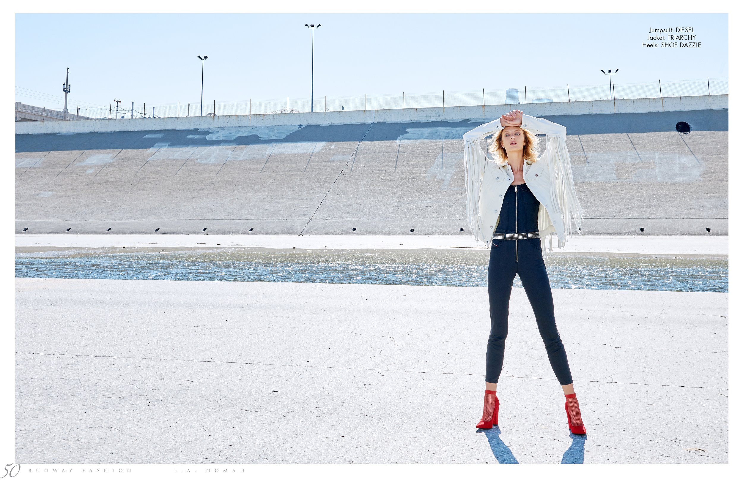 Fashion model standing in an outdoor space with a concrete wall and water feature, wearing a white fringed jacket, black jumpsuit, and red high heels.