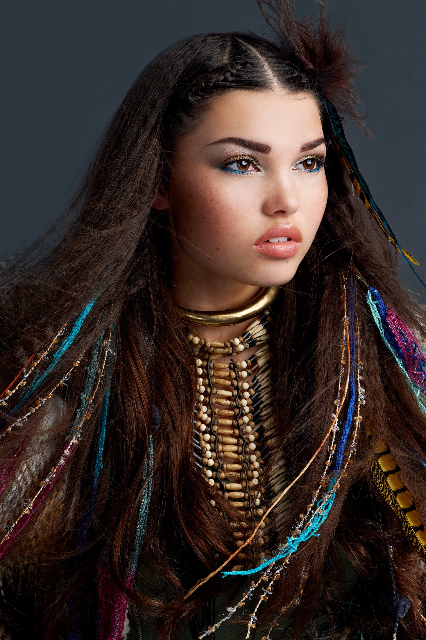 A young woman with long brown hair styled with braids and colorful feathers, wearing native-inspired jewelry including layered necklaces, a choker, and makeup with shimmery eye shadow and nude lipstick, against a dark background.