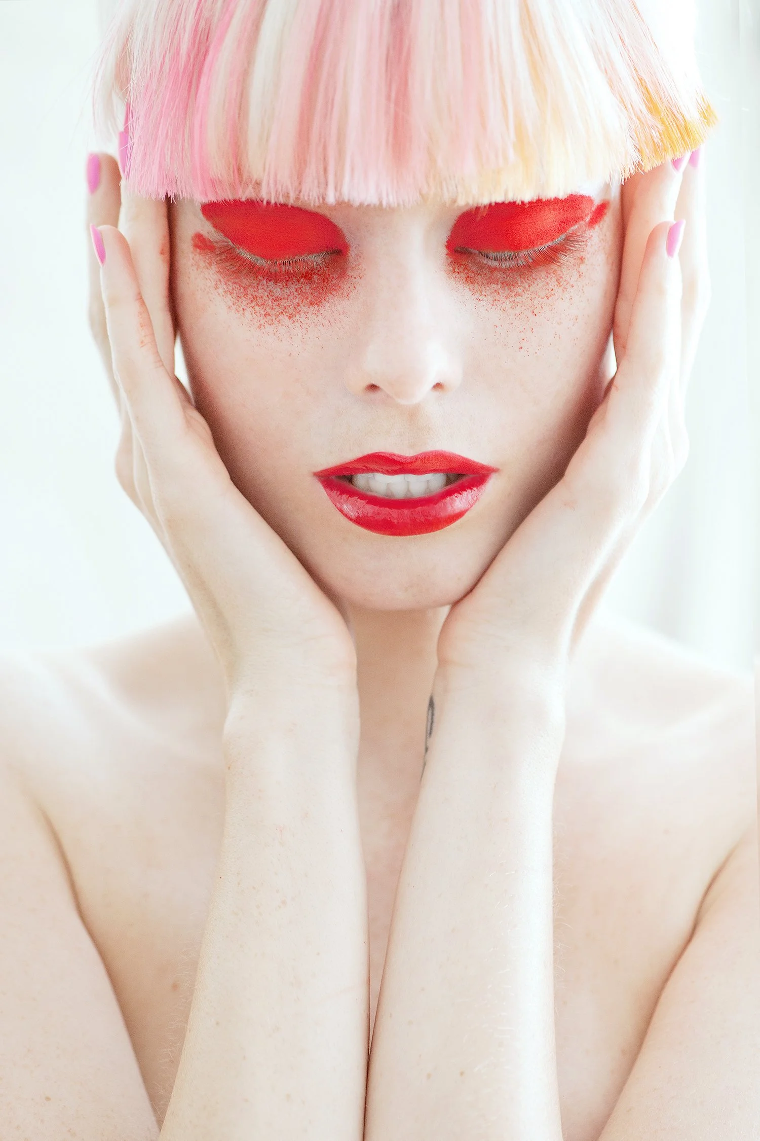A woman with pastel pink and orange hair, with eyes closed, wearing red eyeshadow, lipstick, and nail polish, holding her face with both hands.