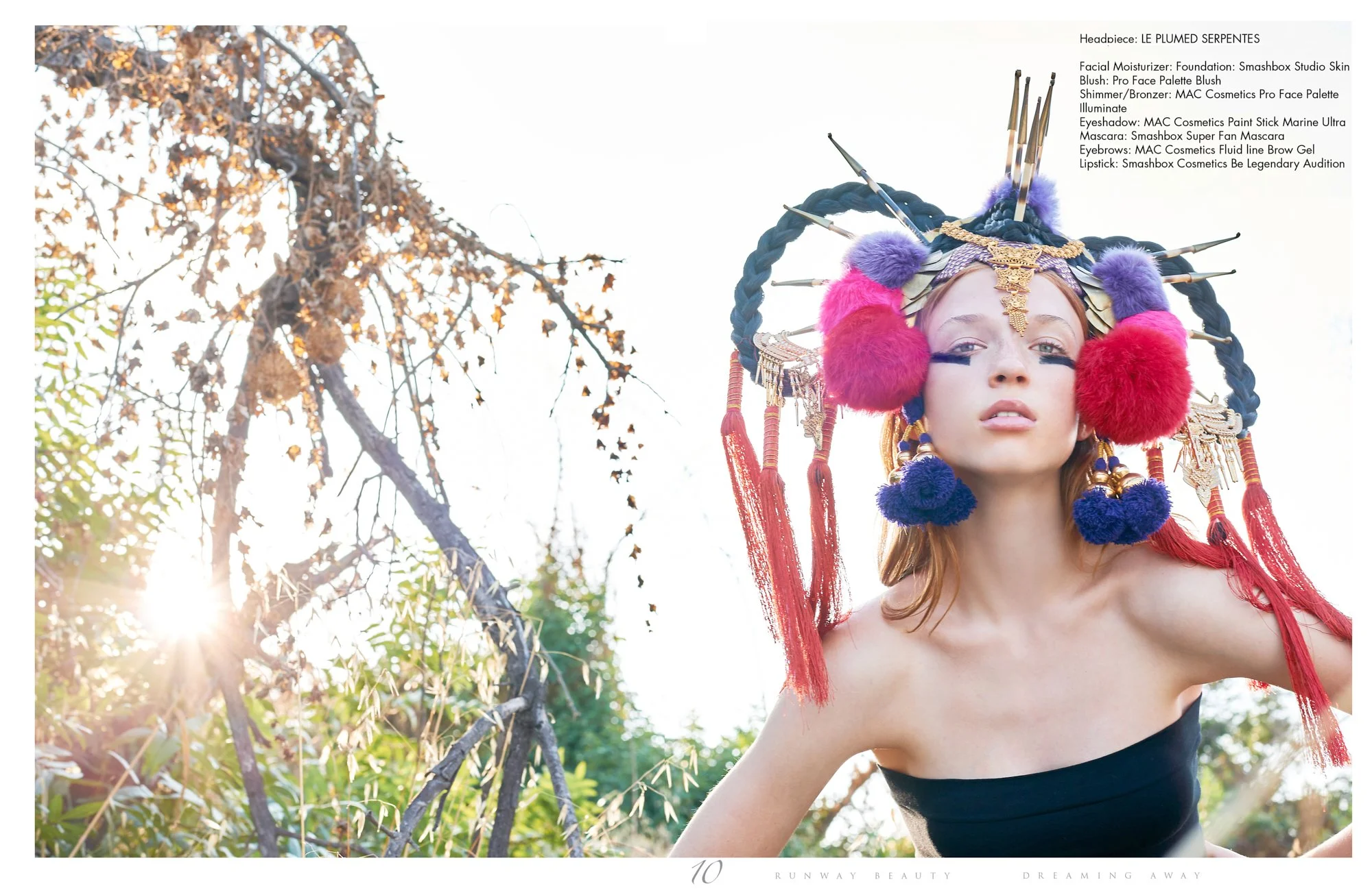 A woman wearing an elaborate headpiece with colorful pom-poms, feathers, and gold accents, posed outdoors with sunlight blooming behind trees in the background.