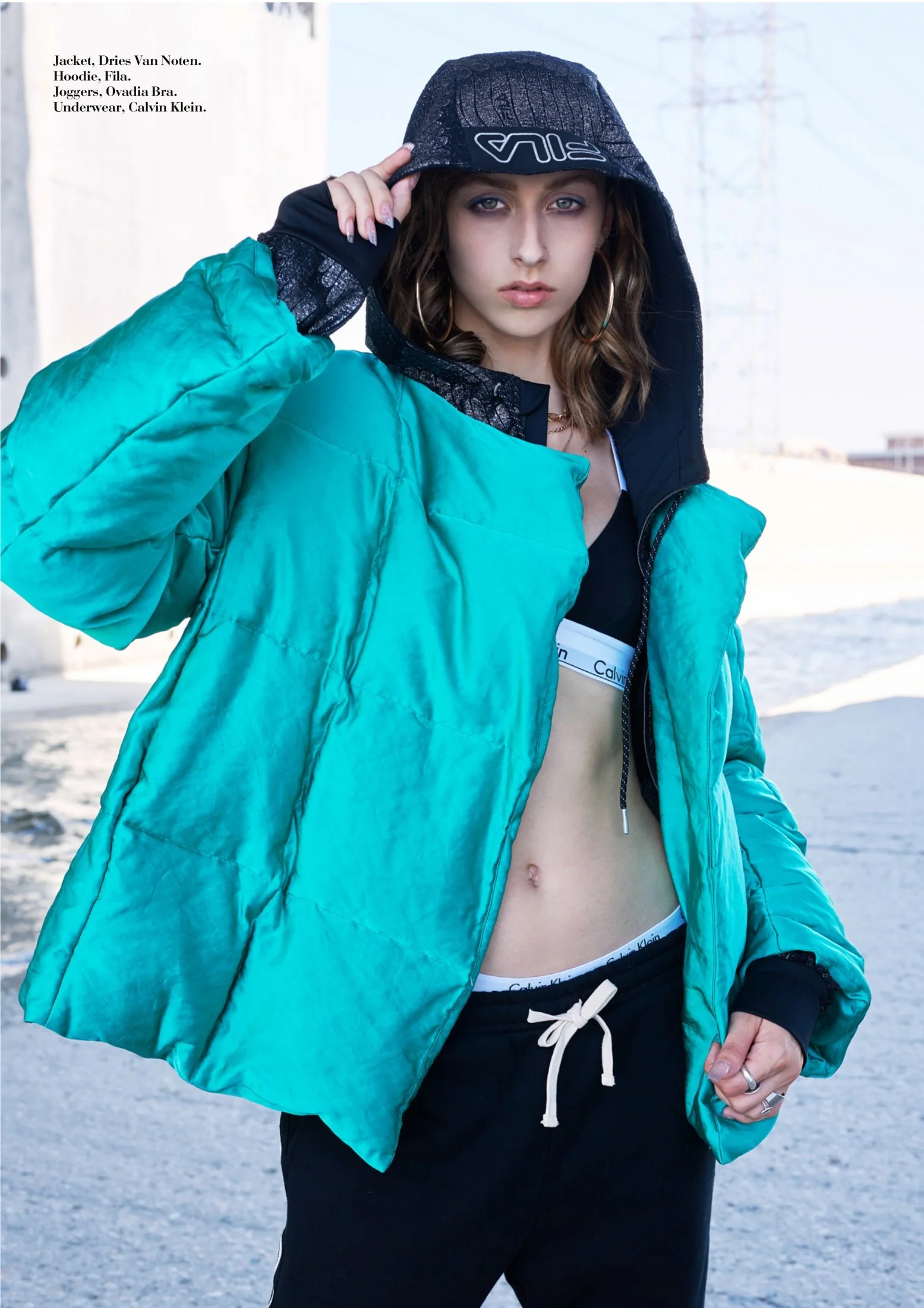 A young woman with wavy brown hair, wearing large hoop earrings, a black hoodie with the hood up, a turquoise puffy jacket, black sweatpants with a white drawstring, and a visible Calvin Klein underwear waistband. She is standing outdoors in an urban