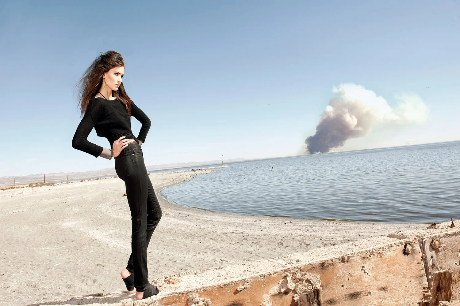Cinematic denim campaign shot by photographer Daniella Hehmann at Salton Sea for Eugene Jeans featuring a model in an all-black ensemble posed against the vast desolate shoreline with smoke in the distance.