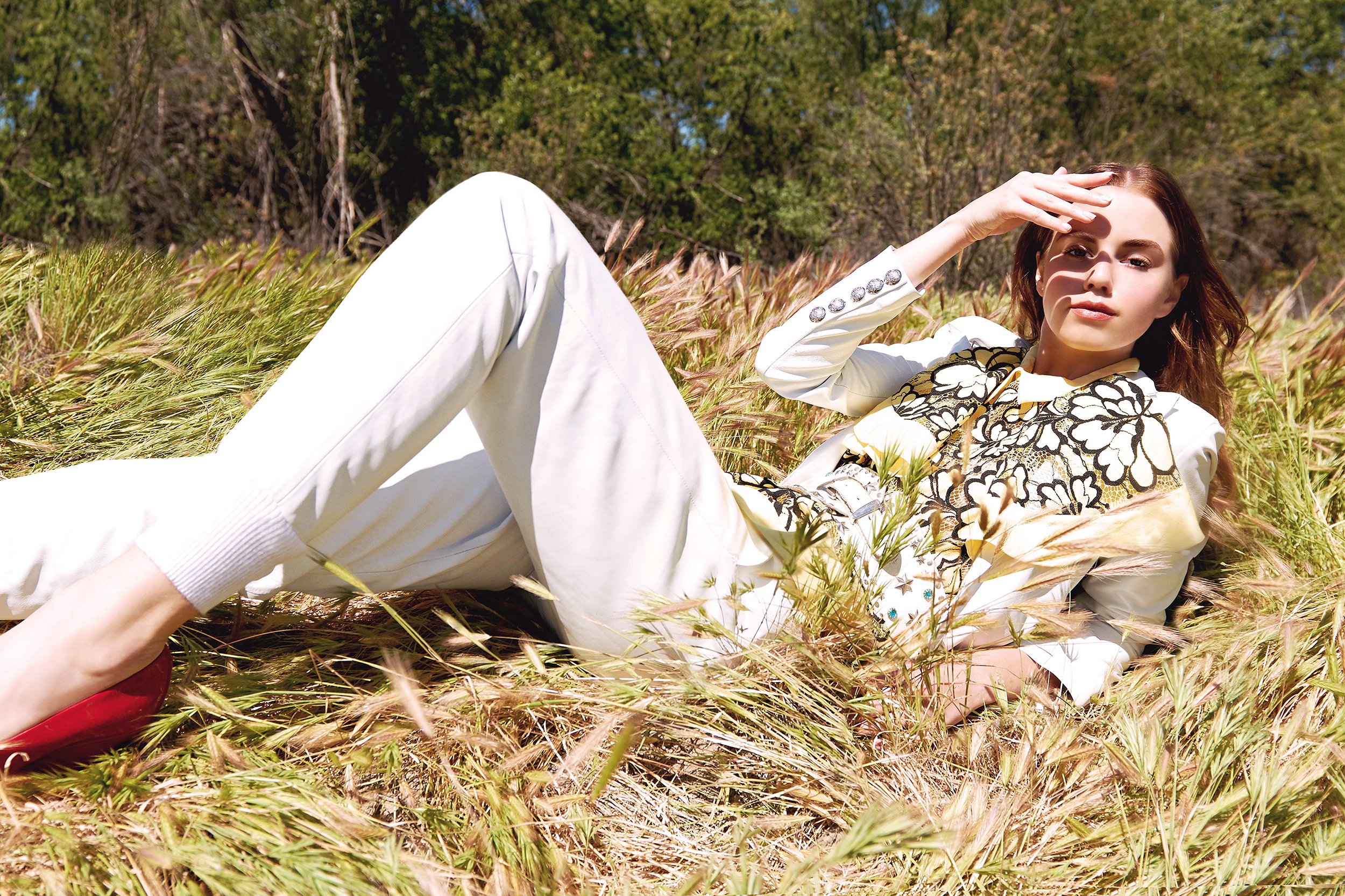 A woman with brown hair lying in a field of tall grass, wearing a white outfit with black and gold embroidery, and red shoes, shading her eyes with her hand.