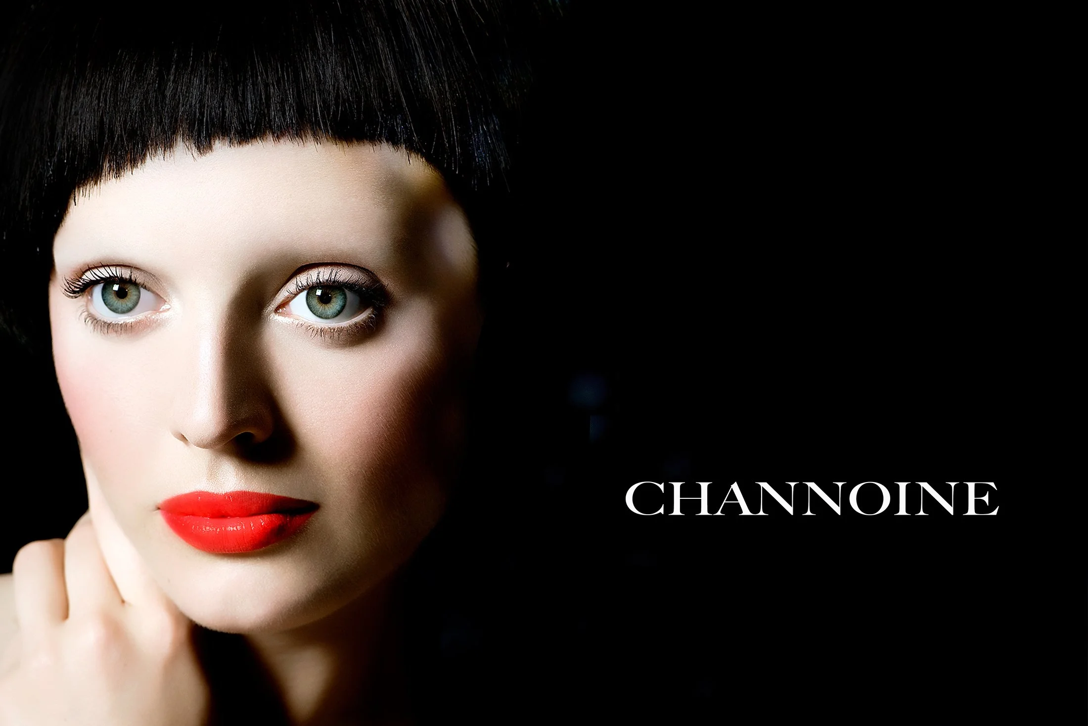 High-contrast advertising photography for Channoine Cosmetics featuring a model with a bold red lip and sharp black bangs. Dramatic, low-key studio lighting creates a sophisticated mood against a deep black background, showcasing professional makeup 