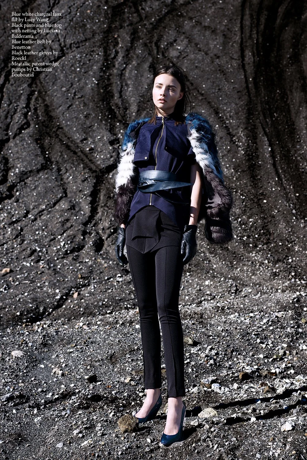 A woman stands outdoors on a dark, rocky terrain. She is dressed in black pants, a navy top with netting, and a faux fur coat with white, blue, and black patches. She wears black leather gloves and black high heels. Her dark hair is styled in a sleek