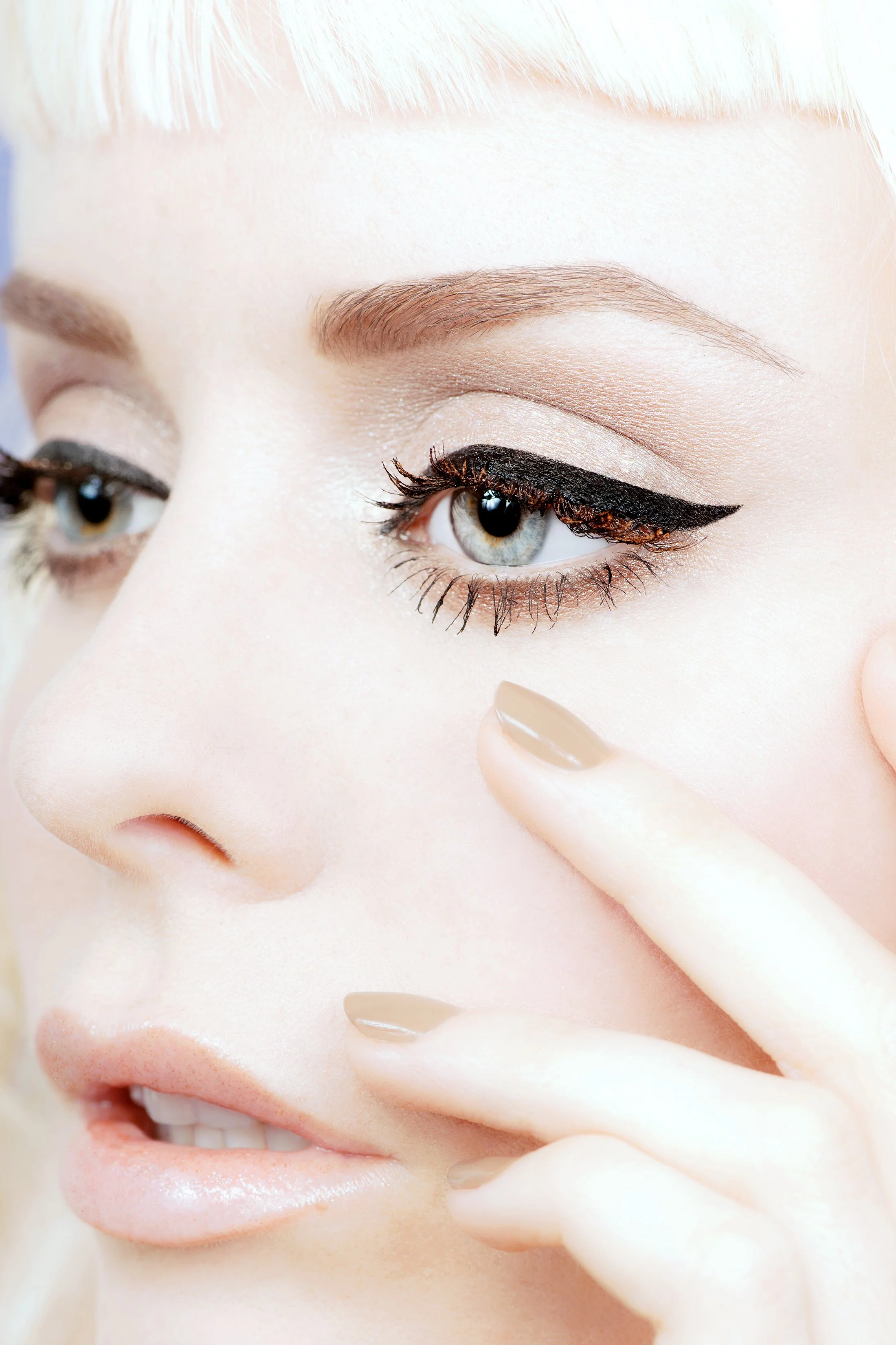 Close-up of a woman with platinum blonde hair, light skin, and makeup, touching her cheek with her index finger, showing her eye makeup and lips.