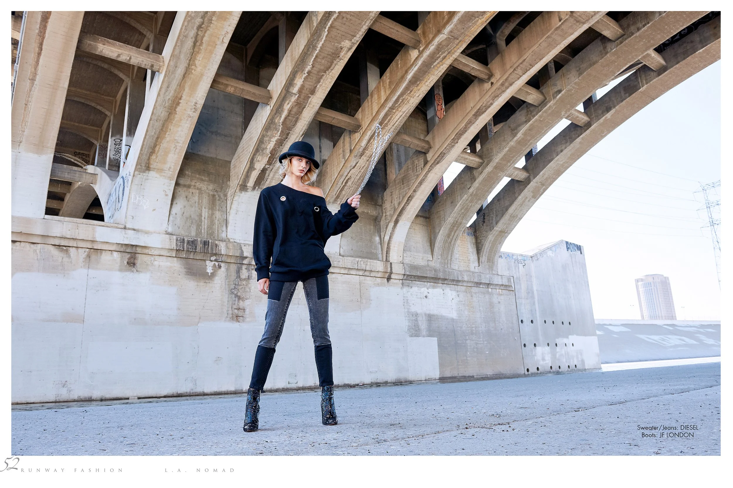 Editorial fashion shot by photographer Daniella Hehmann on location in L.A. The model stands beneath the fourth street bridge, wearing a dark oversized sweatshirt, jeans, and high-heeled boots, with a black hat and holding a chain in her hand, a refe