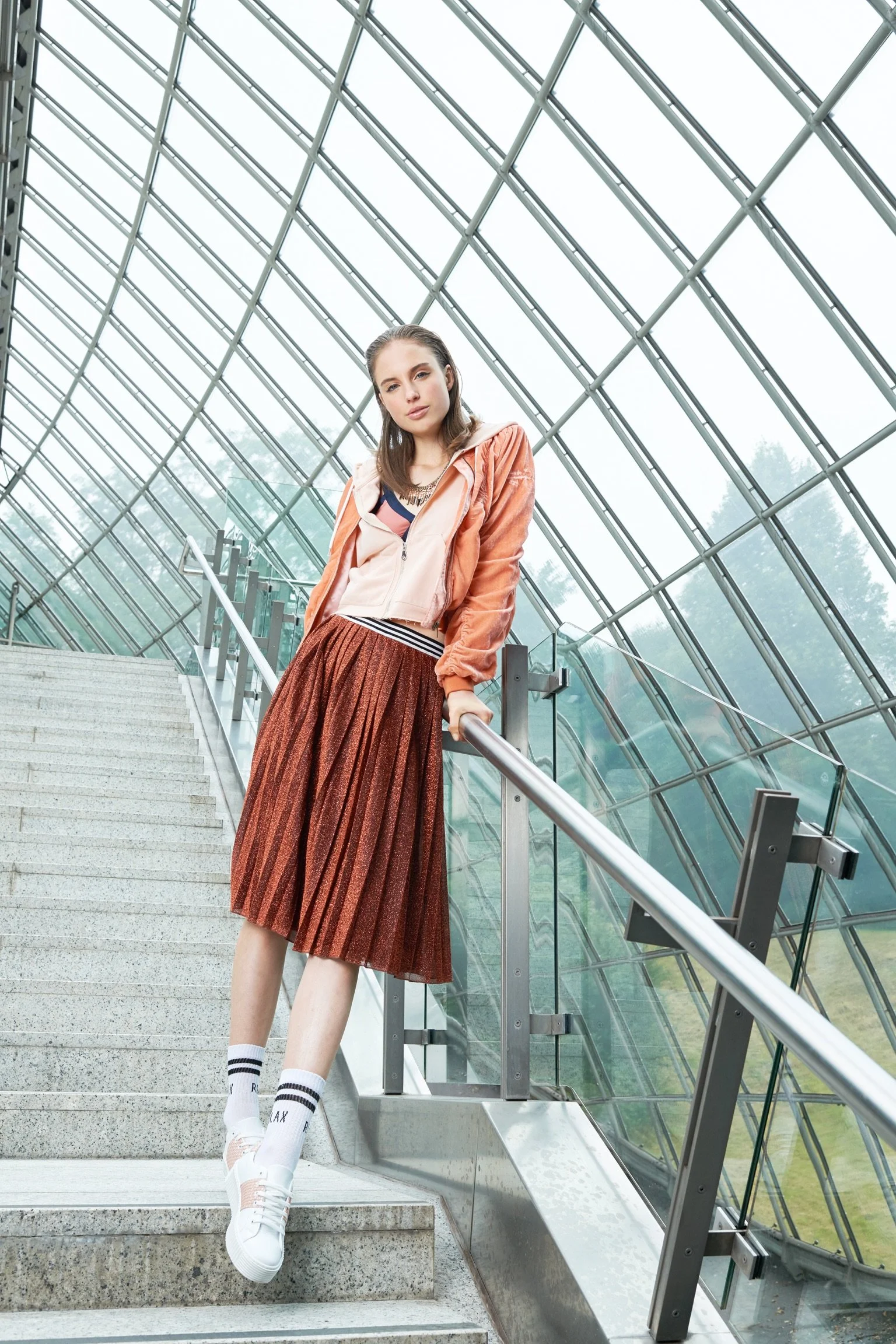 A young woman standing on a staircase inside a modern glass structure, wearing a peach-colored jacket, a striped shirt, a metallic pleated skirt, white sneakers, and socks, with glass walls and metal railings around her.