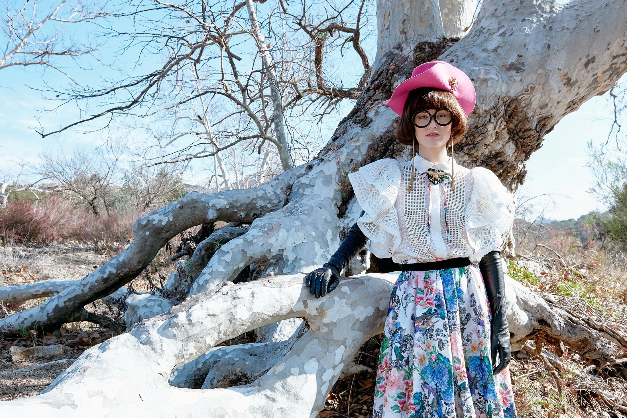 A concluding editorial shot by Daniella Hehmann wearing a pink wide-brimmed hat, black gloves, large black glasses, white lace top, and a long floral skirt, standing outdoors against a large fallen tree with peeling bark and leafless branches.