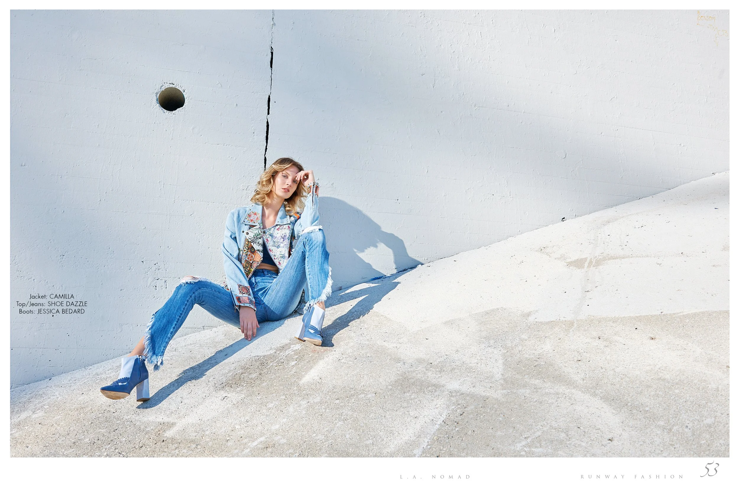 Fashion model sitting on a sandy surface against a white concrete wall, wearing a blue patchwork denim jacket, distressed jeans, and blue high-heeled boots, with text indicating clothing brands and a minimalist runway background.