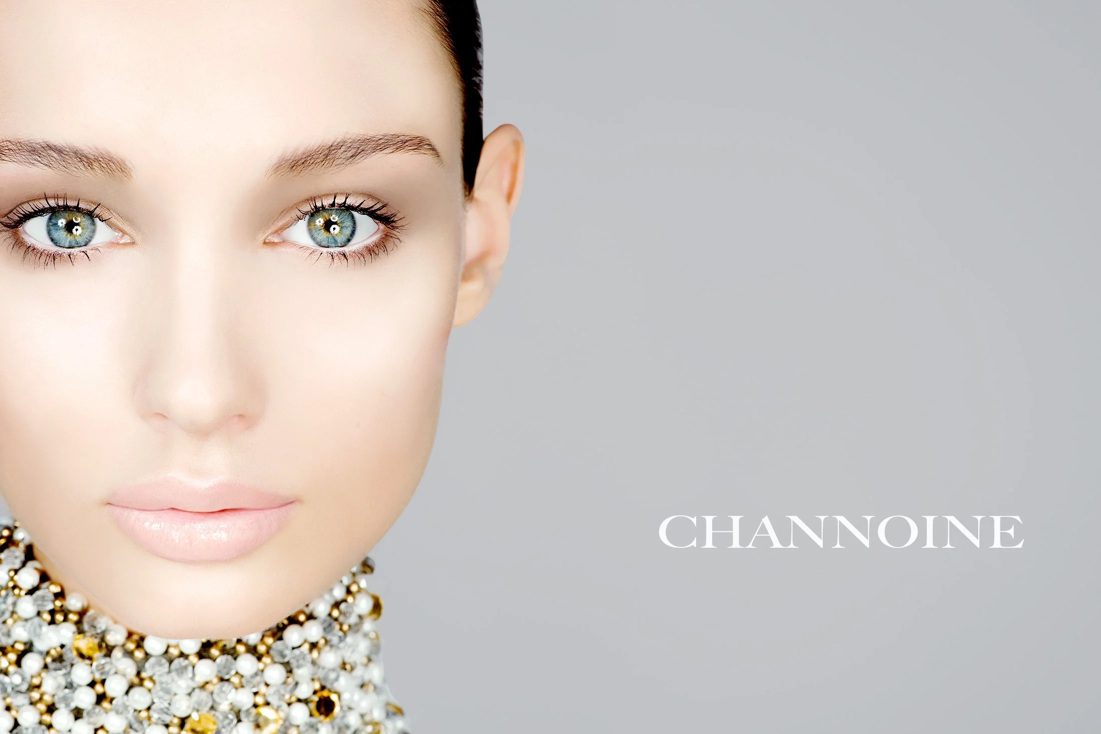 A woman with fair skin, blue eyes, dark hair, wearing pearl and gold jewelry, with the word 'Channoine' written on a plain gray background.