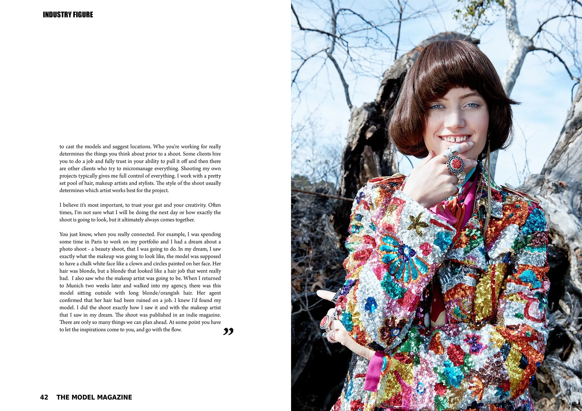 Vibrant fashion portrait by Daniella Hehmann, featuring a model wearing a colorful, sequined jacket with floral patterns and rings, holding a necklace to her lips. In the background, leafless trees and a blue sky are visible.