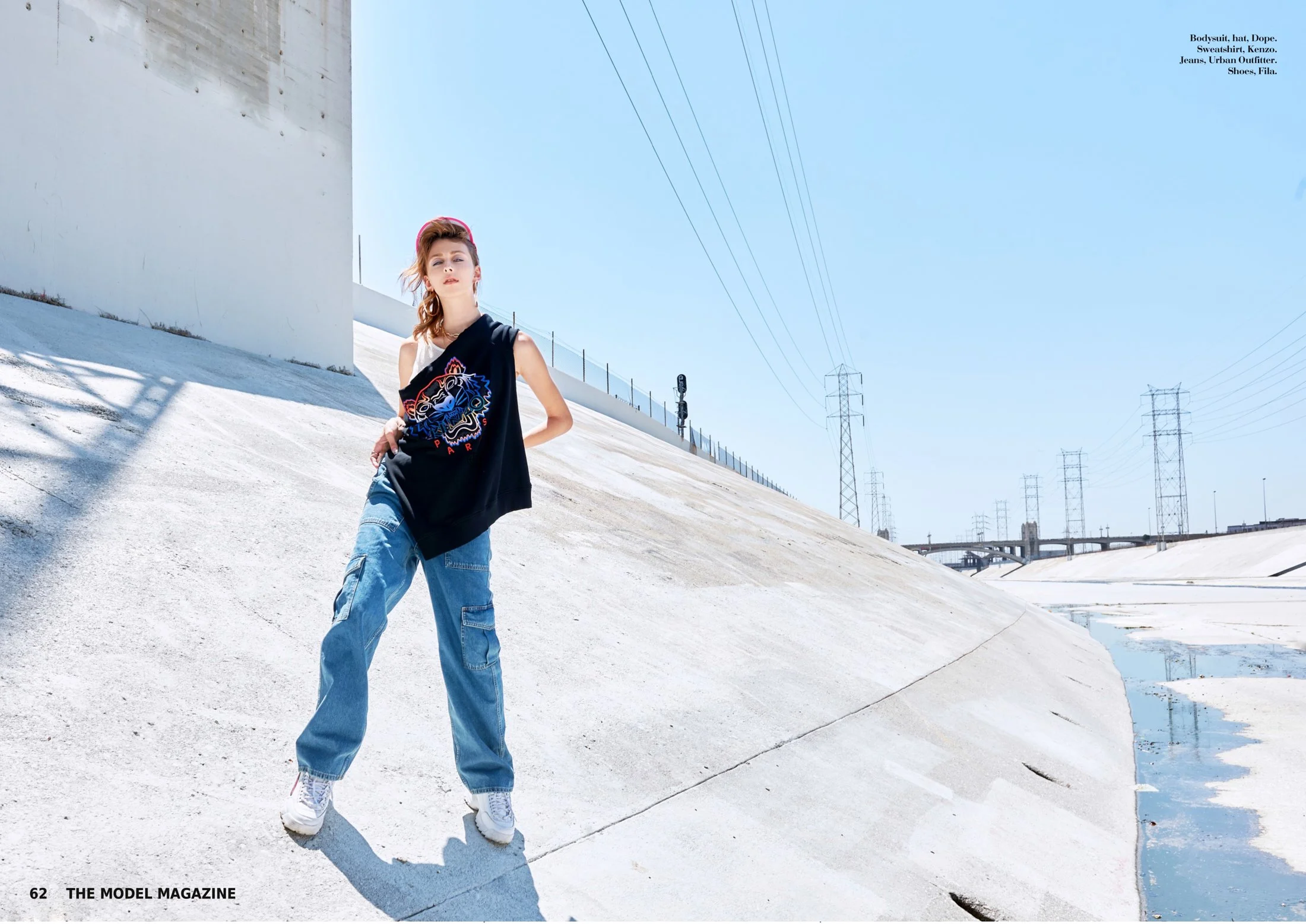 A young woman standing on a concrete slope in an urban setting, wearing a black sleeveless top with a colorful tiger graphic, blue cargo jeans, white sneakers, and a pink headband. The background features electrical pylons, power lines, and a clear b