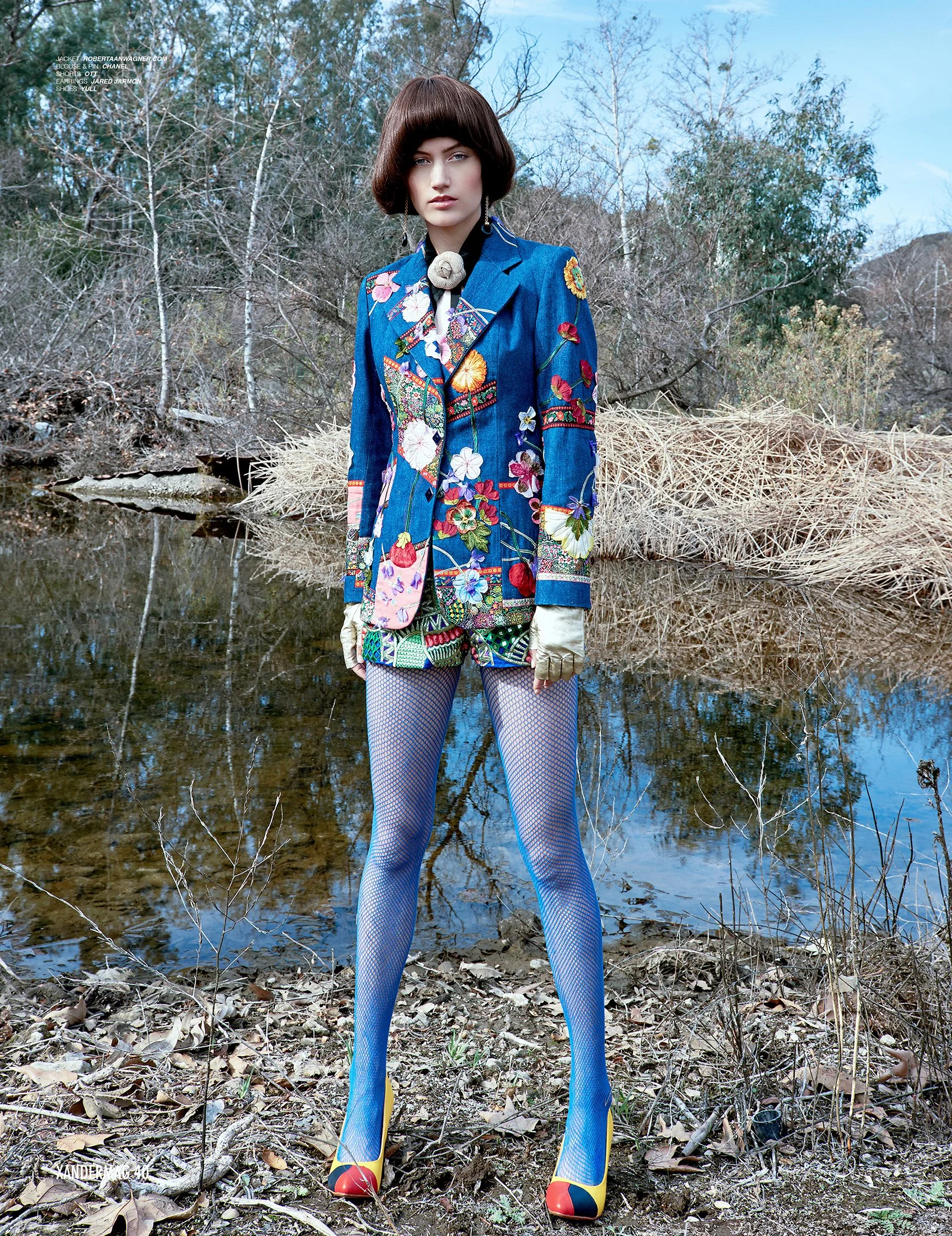 A woman standing outdoors near a creek, wearing a colorful, embroidered denim jacket, patterned shorts, fishnet tights, and heels, with trees and dry grass in the background.