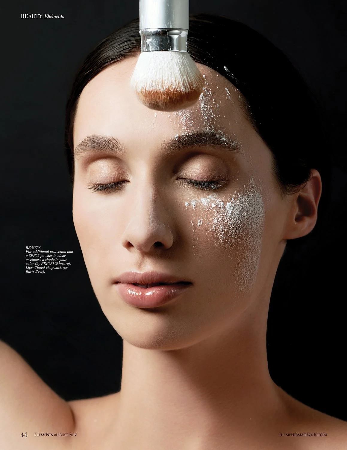 A woman with closed eyes is getting a makeup brush applied to her forehead with white powder. Her face is smooth and her hair is dark and slicked back.