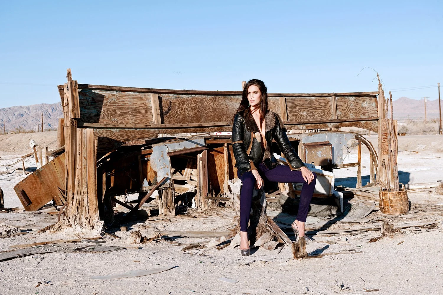 Dramatic fashion shot by photographer Daniella Hehmann at Salton Sea for Eugene Jeans featuring a model in a black leather shearling jacket and purple trousers seated amidst the wreckage of a sun-bleached wooden structure.