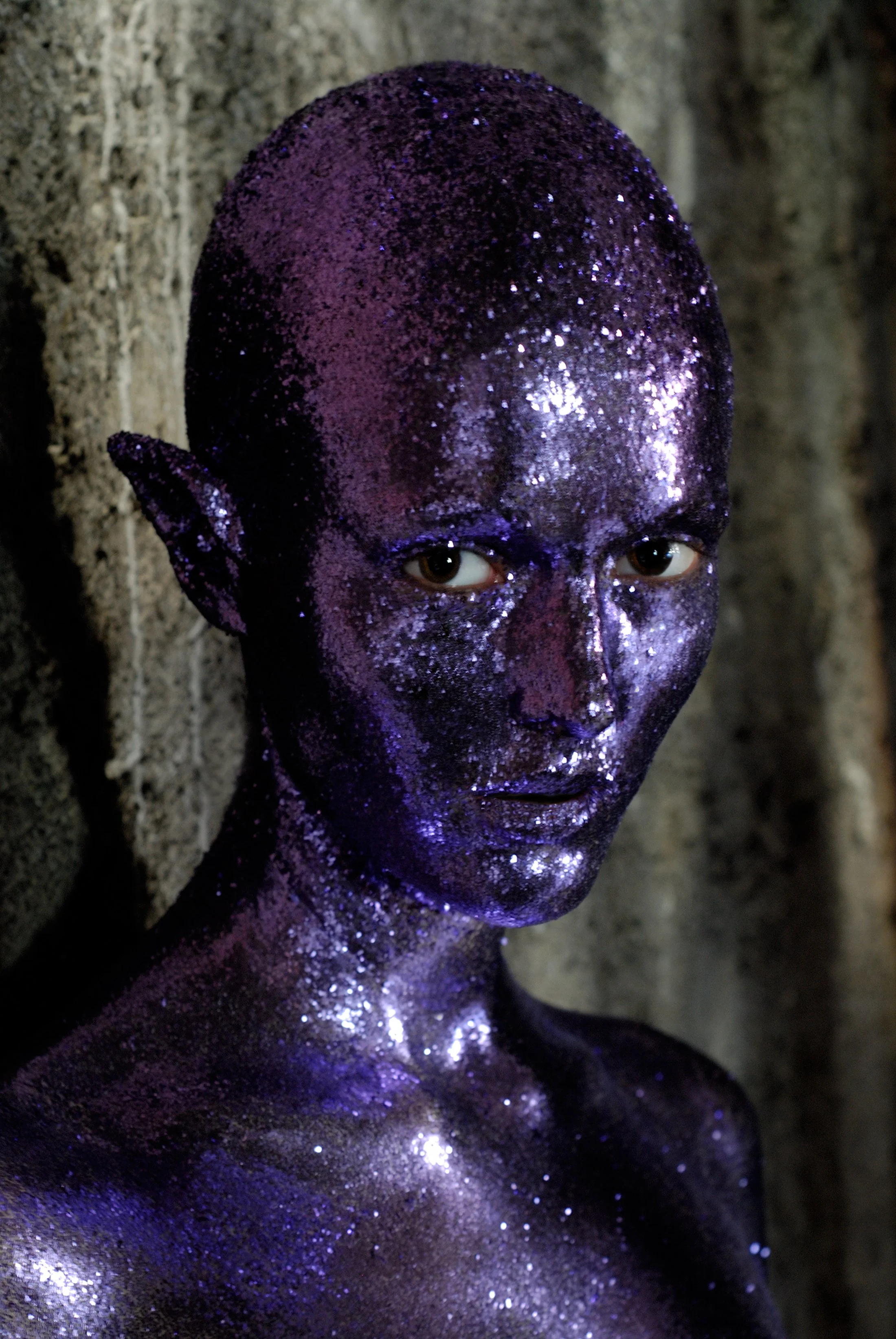 A person with glittering purple body paint and fantasy-like pointed elf ears, with light brown eyes and bald head, standing against a rocky background.