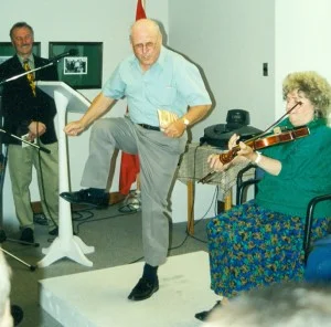 Amand Arsenault kick up his heels while Zélie-Anne fiddles and Georges Arsenault looks on.