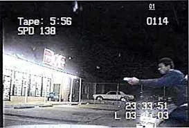The suspect is holding his very shiny cell phone in a two-handed, pointing grip identical to a combat shooting grip. He is pointing (aiming) at Officer Hathorn who has just driven up on the left, out of view