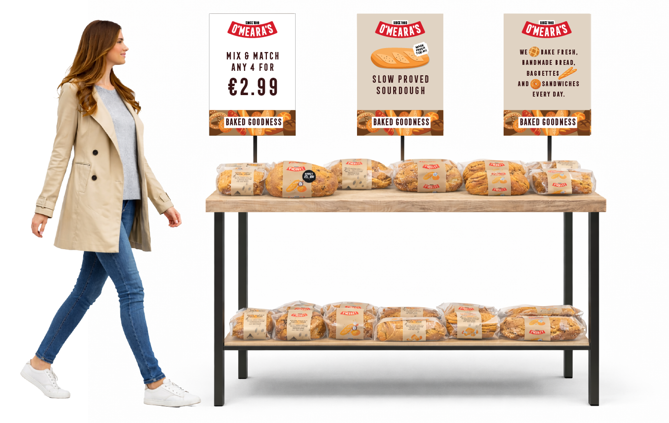 A woman shopping at a bakery stand with various types of bread wrapped in plastic, two signs above the breads advertising deals, baked goods and bakery items, in a store with a light background.