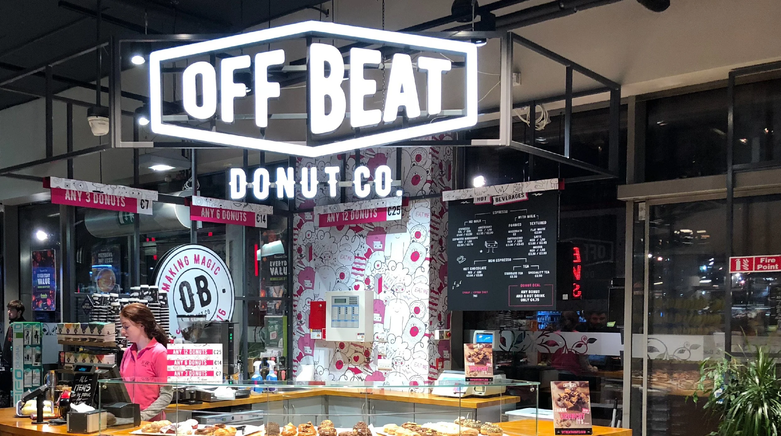 Inside a donut shop called Off Beat Donut Co., with a woman working behind the counter, a display of donuts, and neon signs advertising different donut offers.