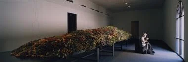 mantle, 1998, Miami Art Museum; a figure, hand-sewing wool coats, sits in front of a long steel table where multiple speakers connected to seven short wave radios are buried in a bed of fresh-cut flowers; photo courtesy Thibault Jeanson