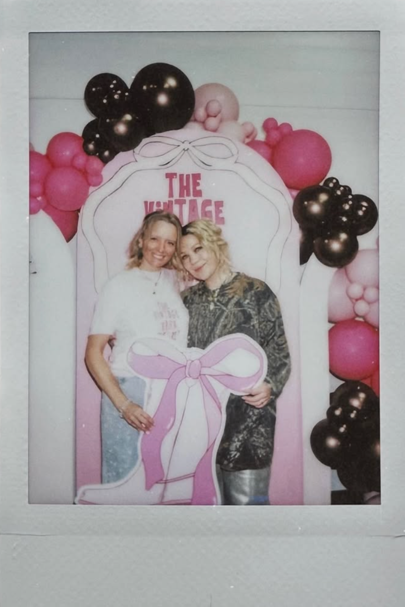 Two women posing in front of a decorative backdrop with pink, black, and beige balloons, and a sign that reads 'The Vintage'. They are smiling and standing close together, with one woman wearing a white t-shirt and jeans, and the other in a dark, patterned shirt.