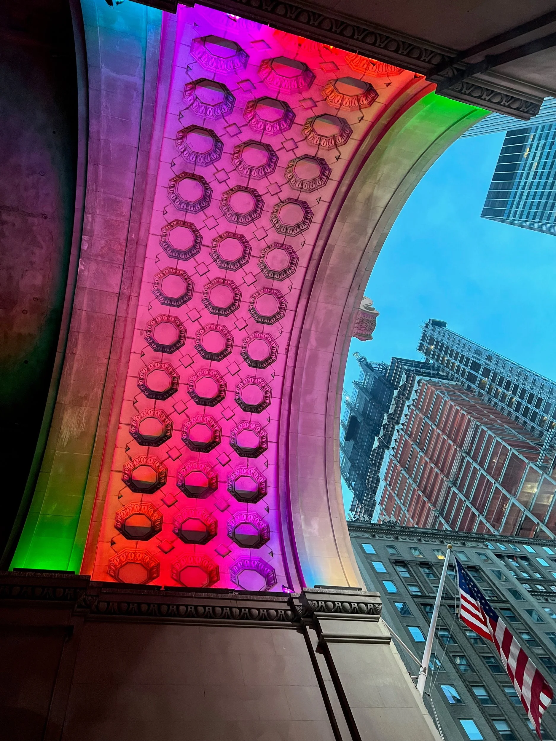 Ornate arch lit in rainbow colors, seen from below against city towers