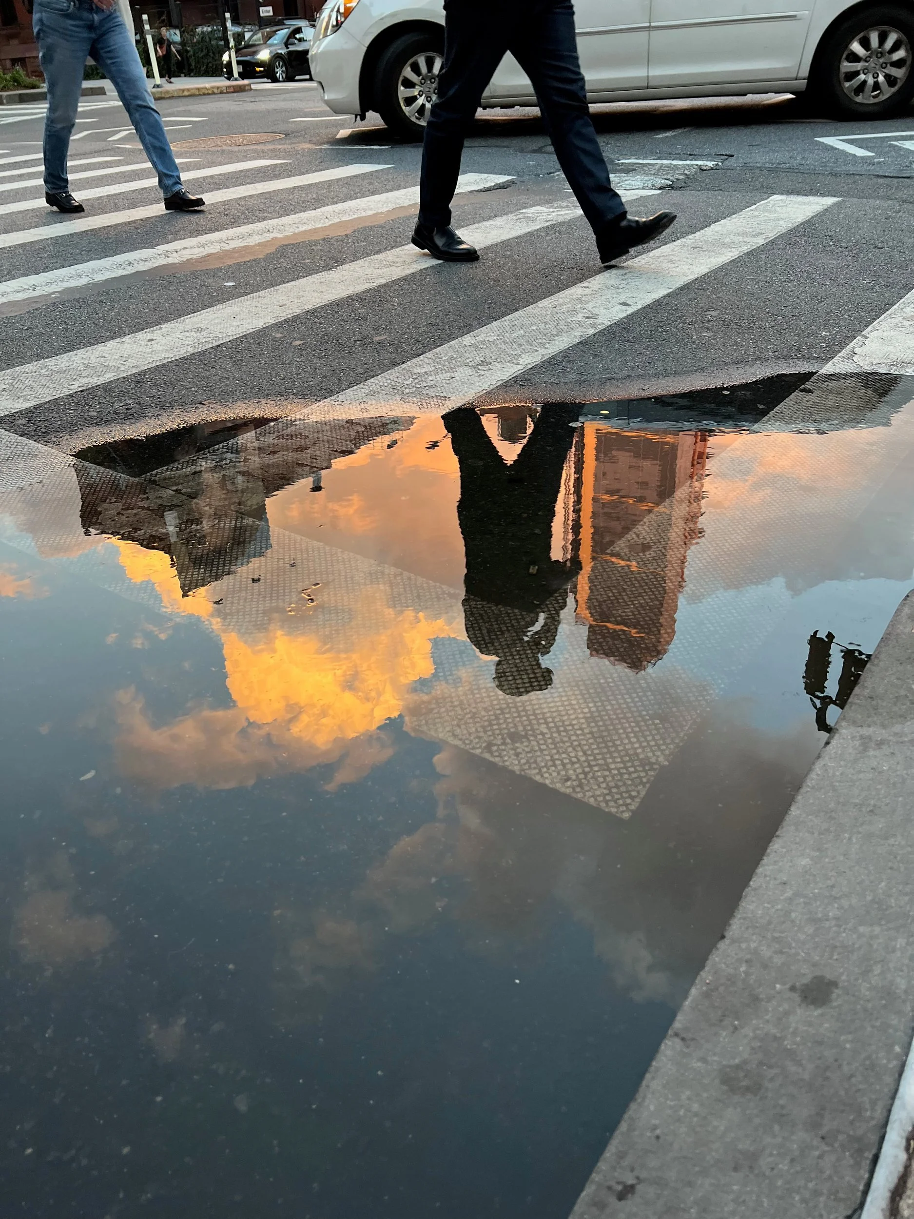 Pedestrians and sunset clouds reflected in a crosswalk puddle, New York City